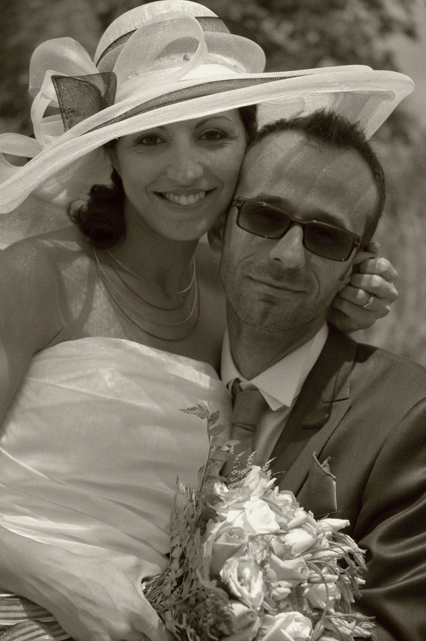 Una coppia di persone sorridenti, donna con cappello grande e uomo con occhiali da sole, si abbracciano con bouquet di fiori, in un contesto esterno.