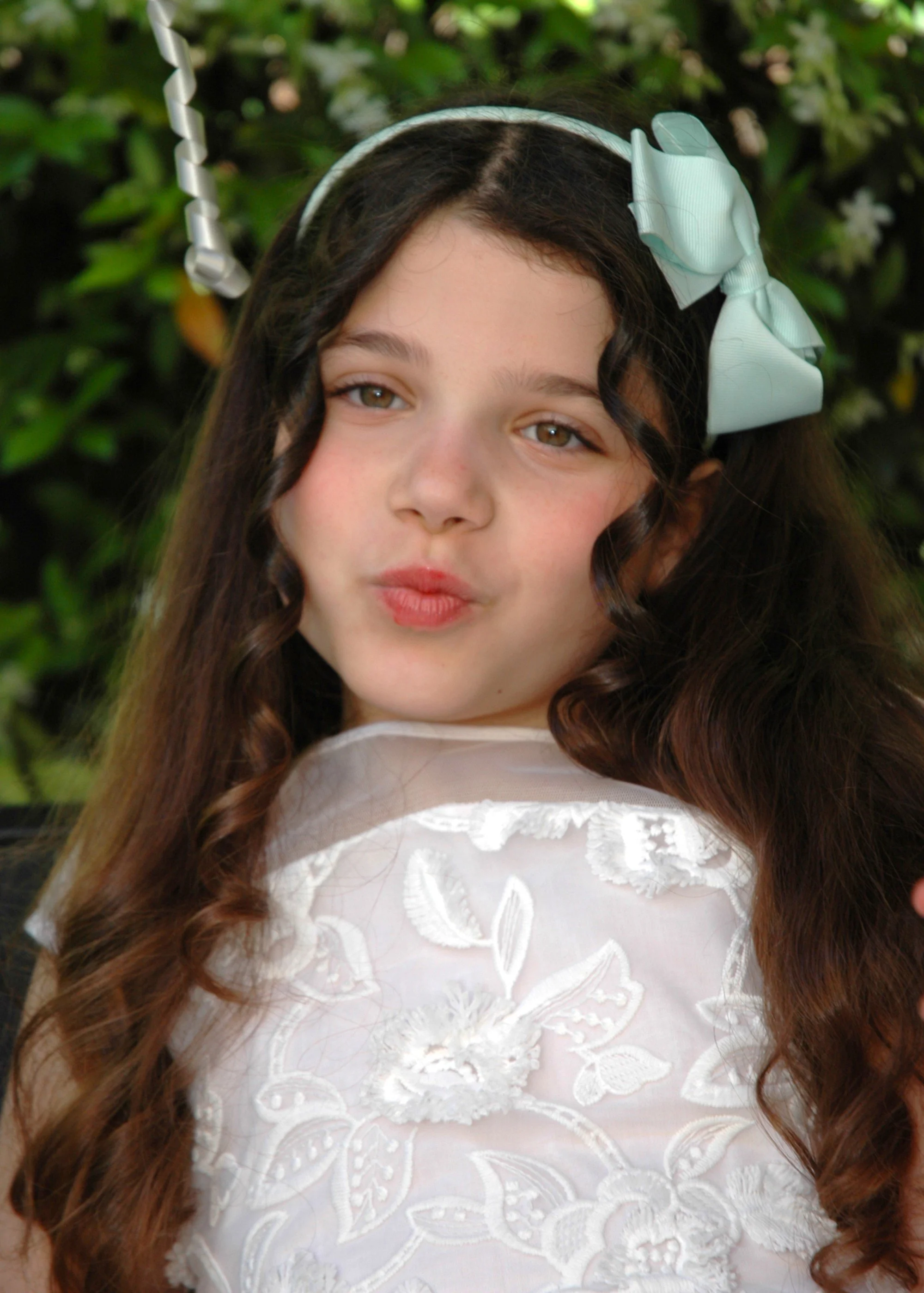 Ragazza con lunghi capelli mossi, indossa un fermaglio con nastro azzurro e un abito bianco ricamato con motivi floreali.