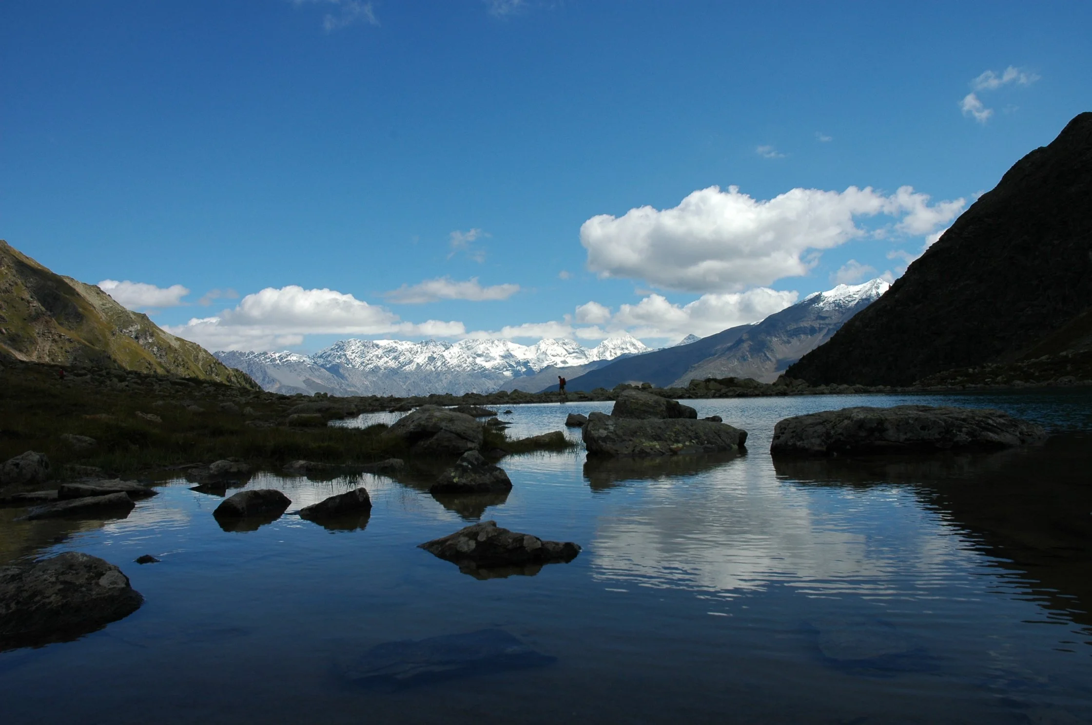 Paesaggio montano con acque serene, montagne innevate sullo sfondo, cielo blu con nuvole sparse e una persona che cammina lungo il fiume.