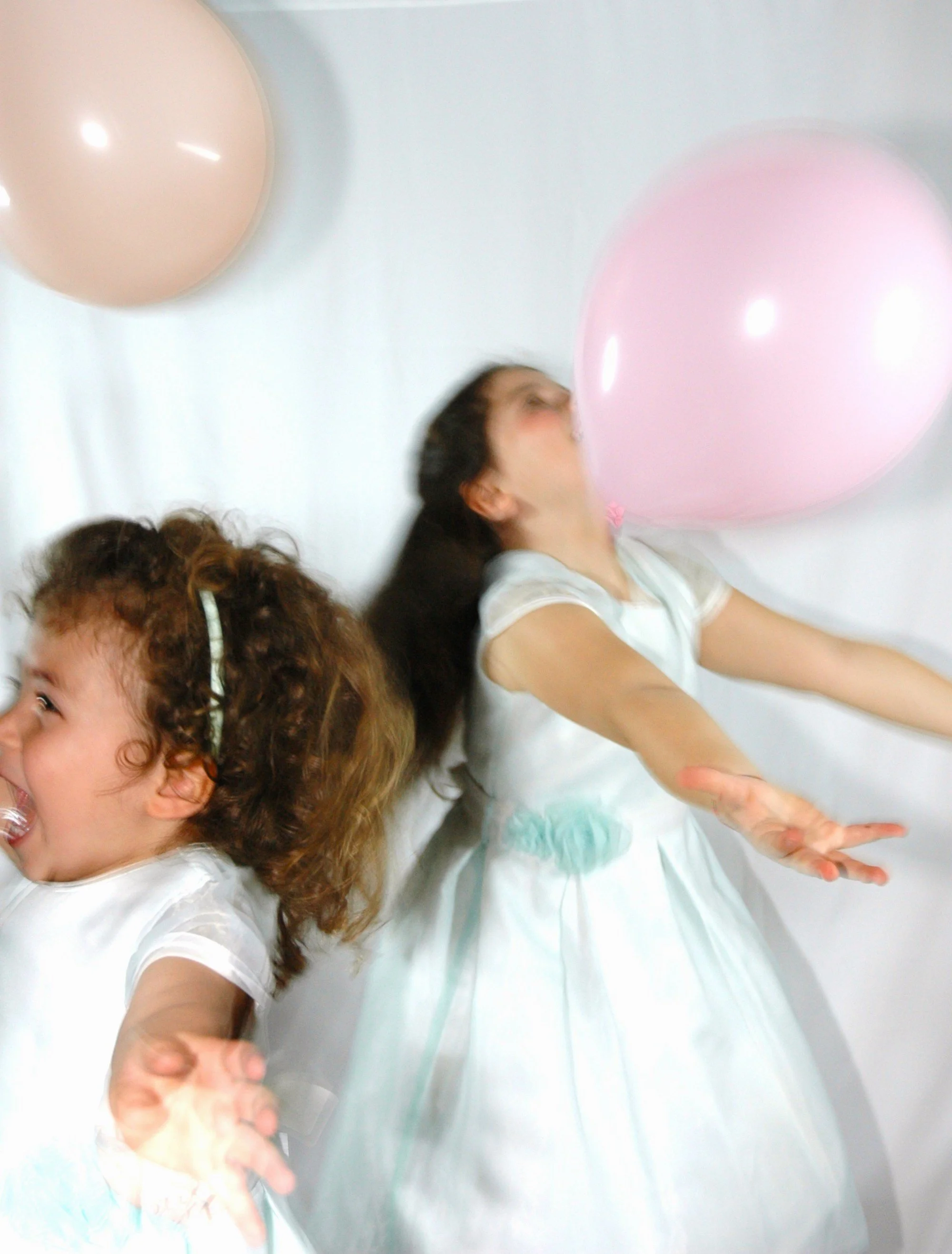Due persone, probabilmente due bambine, che si trovano sul pavimento a terra in una stanza bianca con palloncini rosa e beige, sembrano giocare o festeggiare.