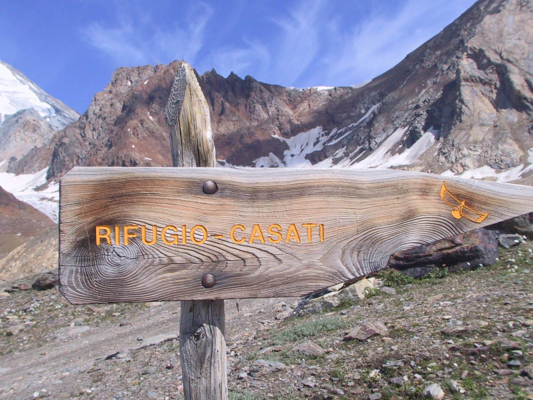 Pannello di legno con scritta in italiano 'RIFUGIO-CASATI' e simbolo di un ventaglio, situato in un'area montuosa con piccole nevi e montagne rocciose sullo sfondo
