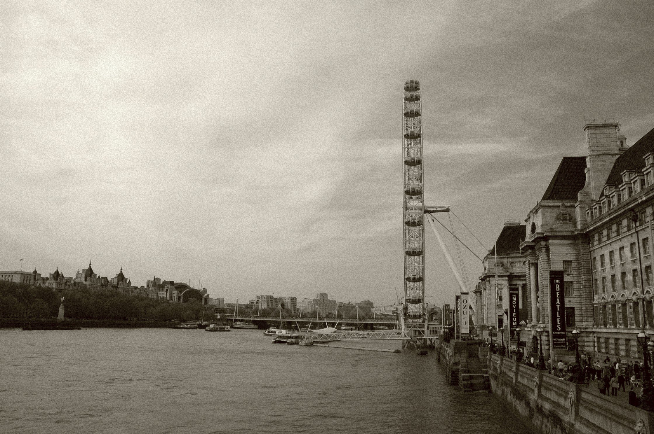 Veduta di Londra con ruota panoramica, edifici storici lungo il fiume e barche sull'acqua in una giornata nuvolosa.