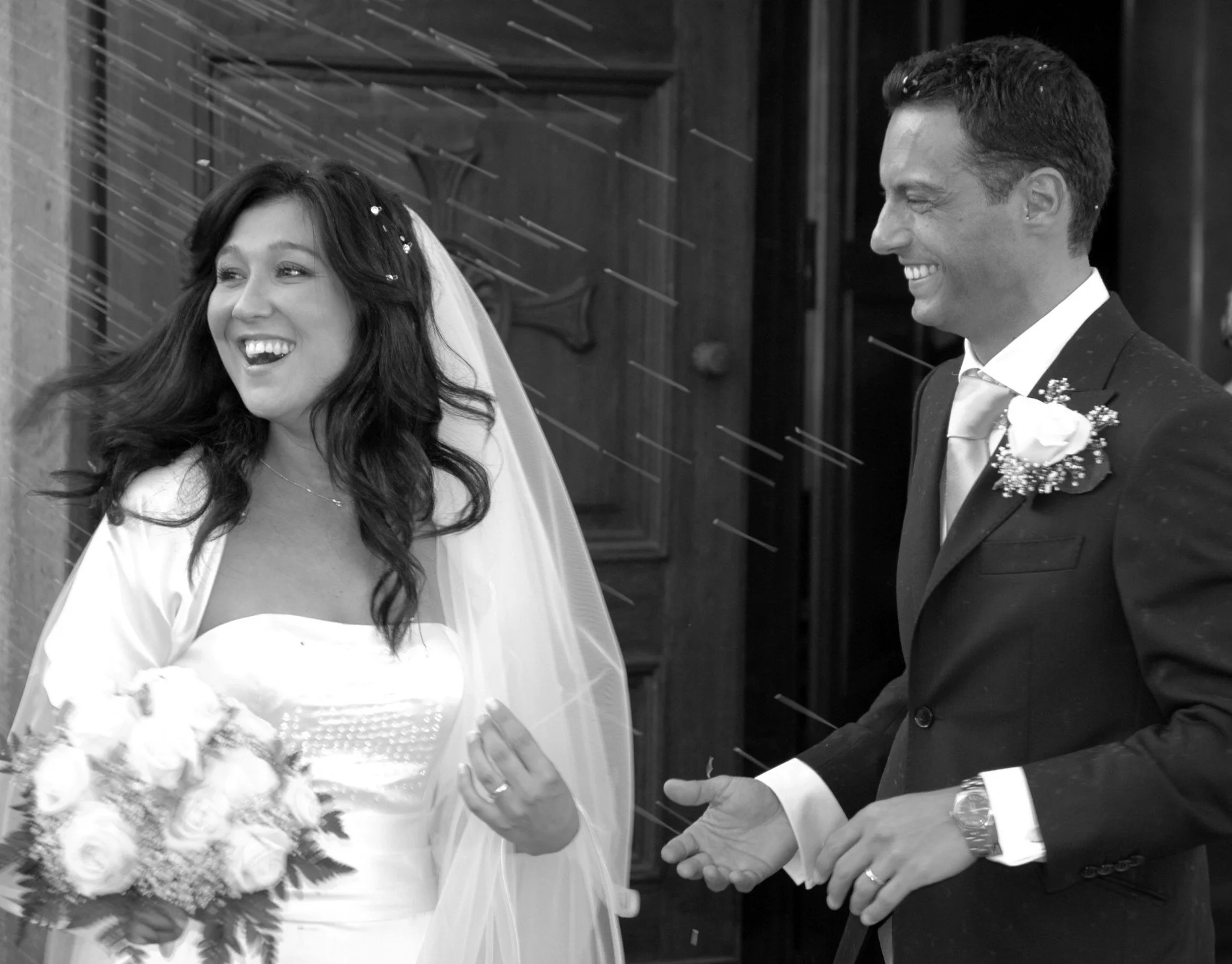 Una coppia di sposi sorridenti durante il matrimonio, la sposa con un vestito bianco e un velo, il marito con uno smoking, intento a scambiarsi un anello.