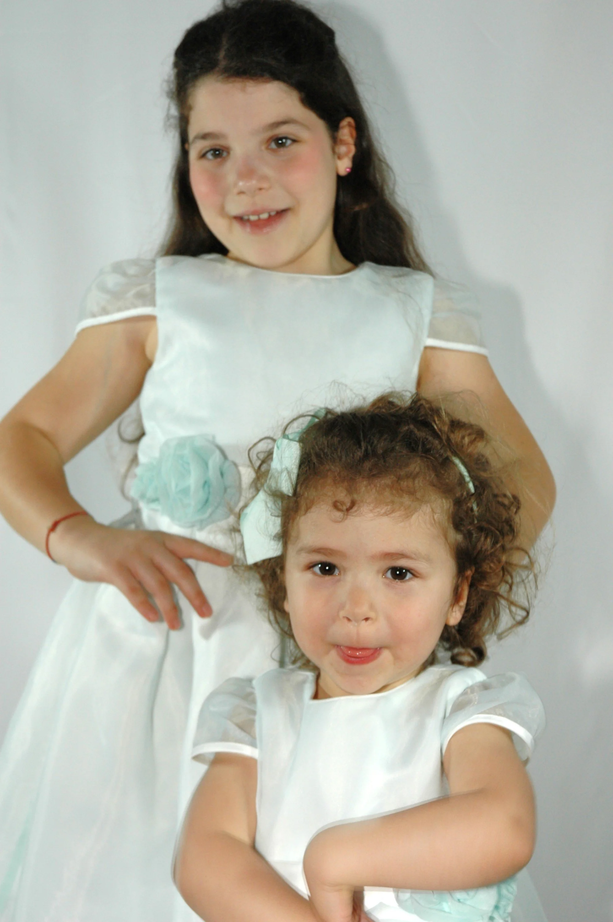 Due giovani ragazze con capelli castani e occhi chiari indossano vestiti bianchi e sorridono, con un muro bianco sullo sfondo. La più giovane in primo piano mostra la lingua, mentre la più grande sta dietro di lei, con le braccia incrociate.