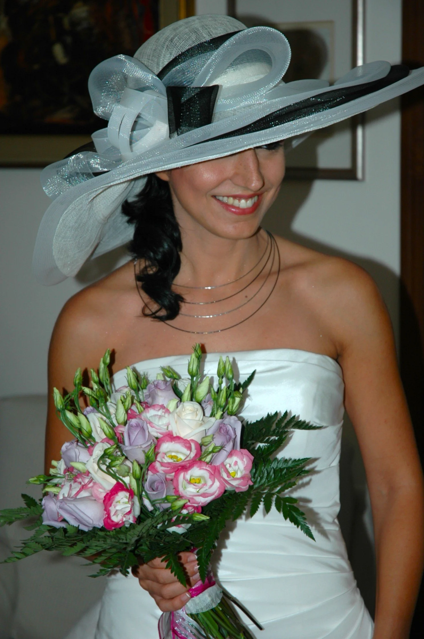 Una donna che indossa un cappello grande e decorato, sorride mentre tiene un bouquet di fiori colorati, indossa un abito bianco da matrimonio.