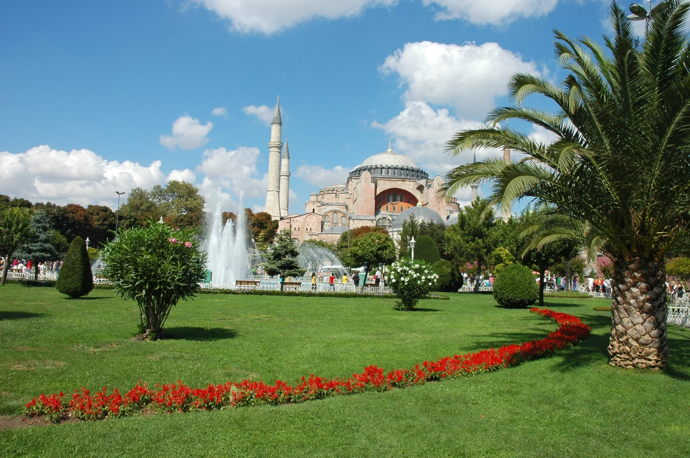 Un parco verde con palme, alberi e aiuole di fiori, fontane e molte persone, con un grande edificio storico con cupola e torri, il tutto sotto un cielo di nuvole.