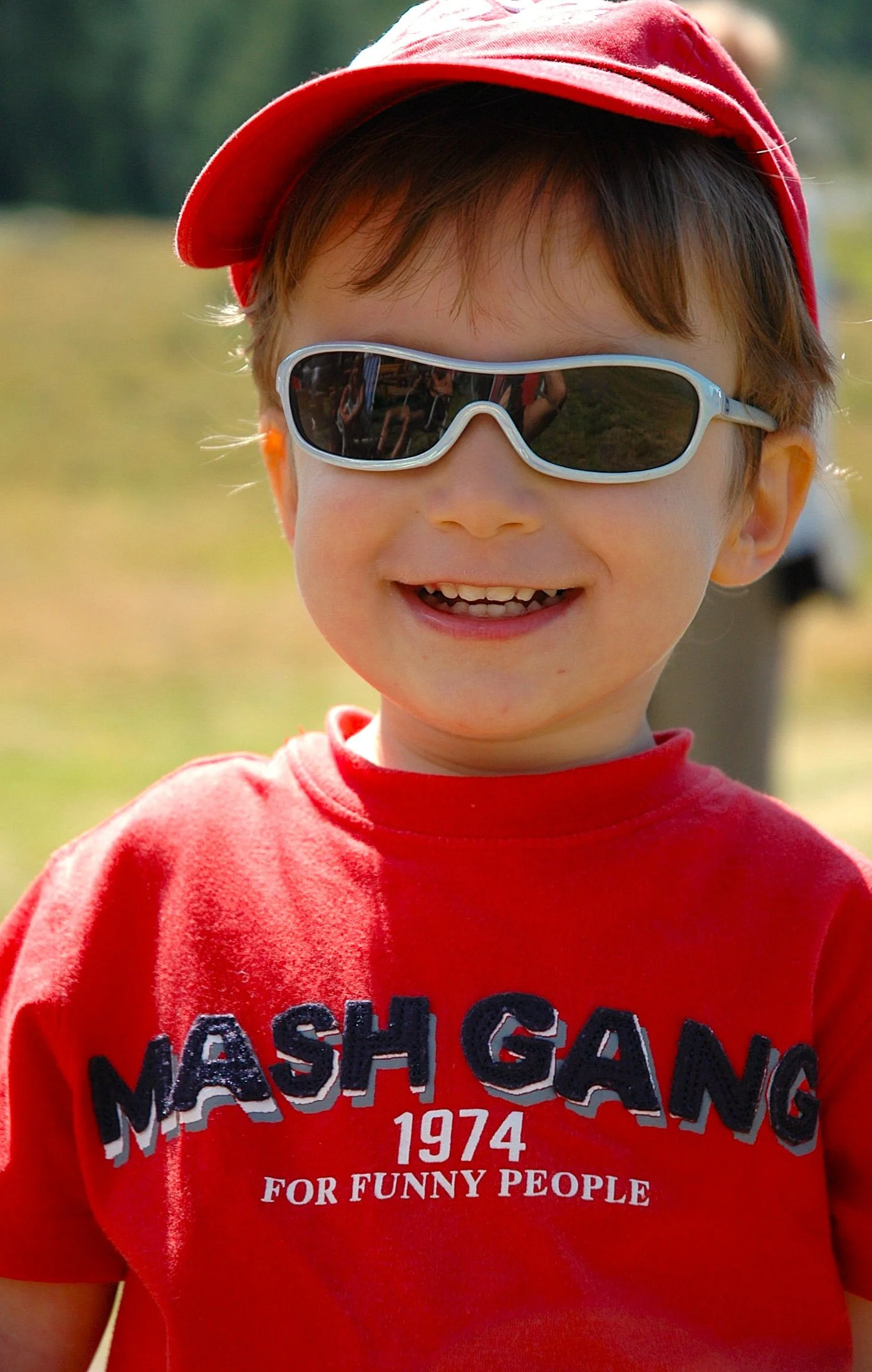 Ragazzo con cappello rosso e occhiali da sole, sorridente, maglietta rossa con scritte nere e bianche.