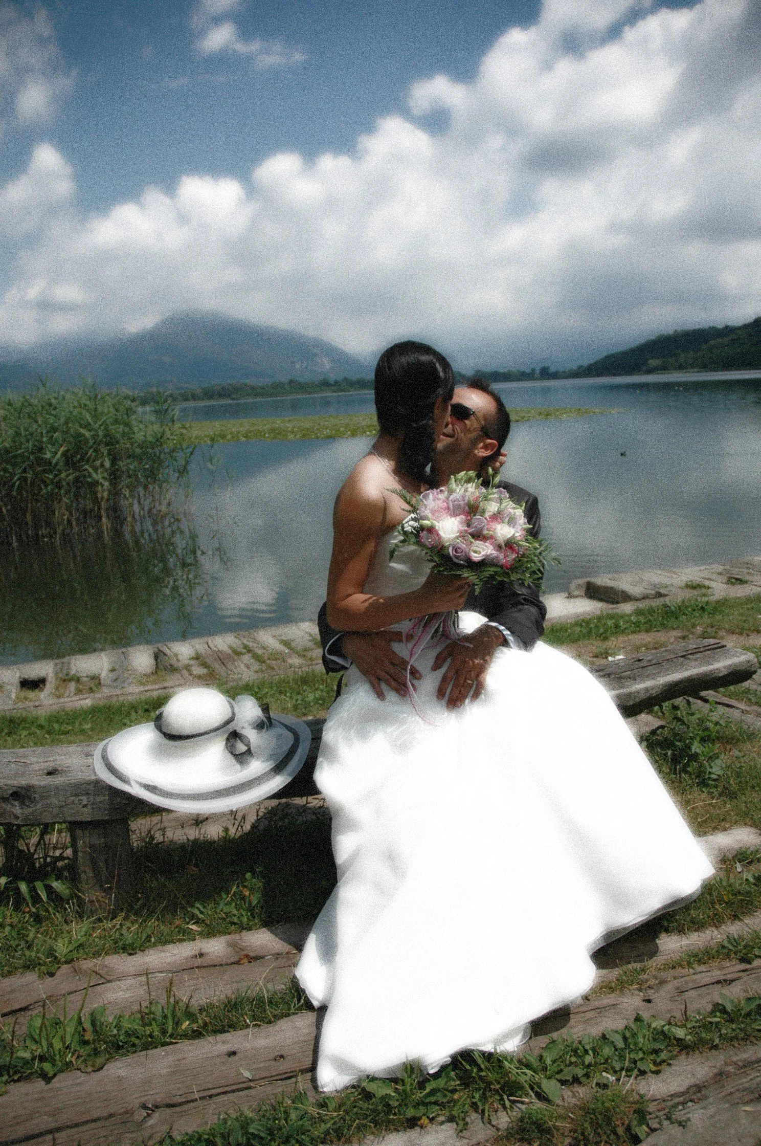Una coppia di sposi si bacia vicino a un lago, con una sposa in abito lungo bianco e un uomo in abito nero. La sposa tiene un bouquet di fiori e c'è un cappello bianco appoggiato su una panchina di legno.