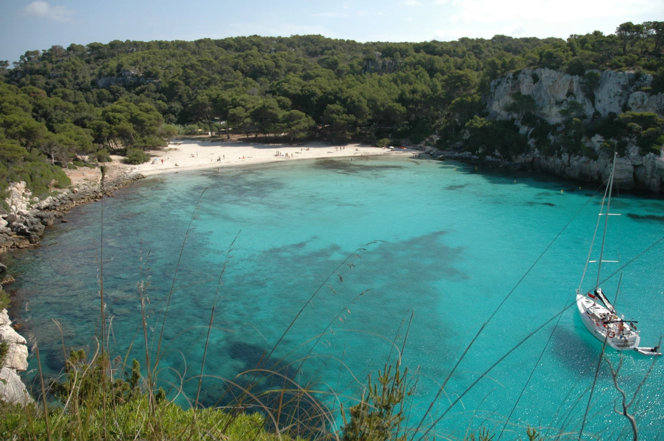 Spiaggia nascosta tra le scogliere con acqua turchese, alcune persone relax sulla spiaggia e una barca ancorata vicino alla riva, circondata da vegetazione verde.