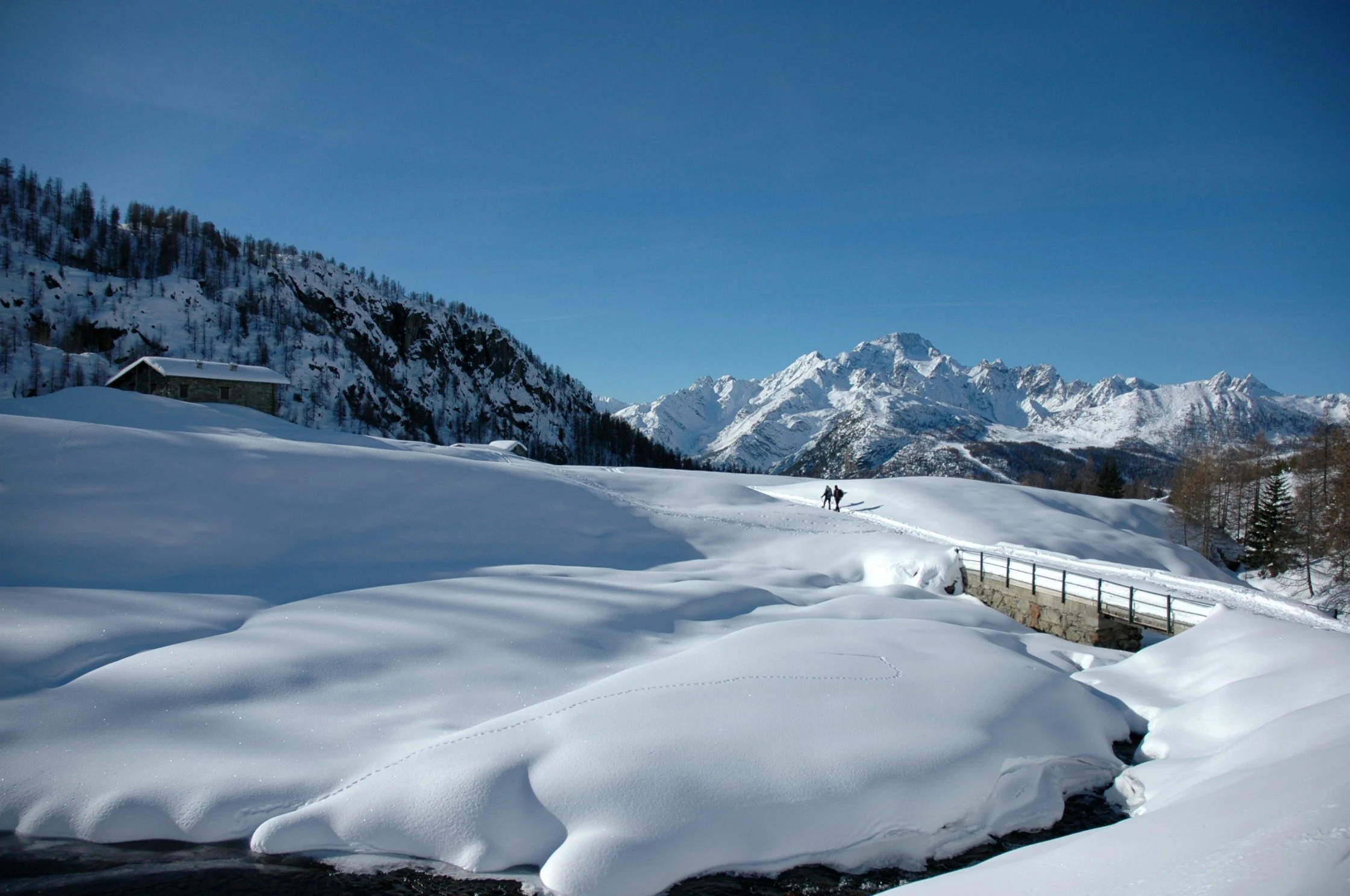 Paesaggio invernale con montagne innevate, una baita, due persone che camminano nella neve e un cielo chiaro.