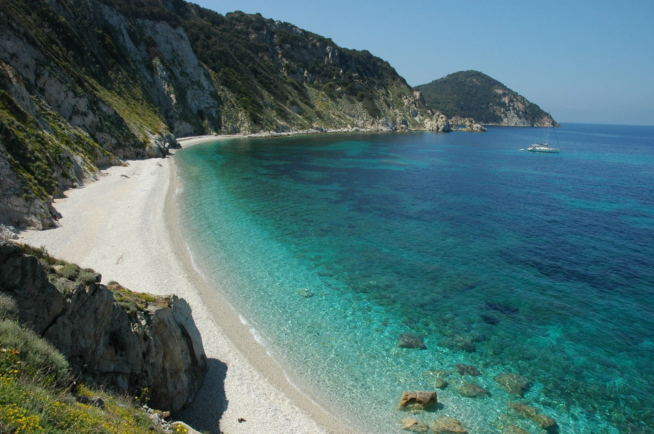 Spiaggia di piccole dimensioni con sabbia chiara e acque cristalline, circondata da alte scogliere verdi.