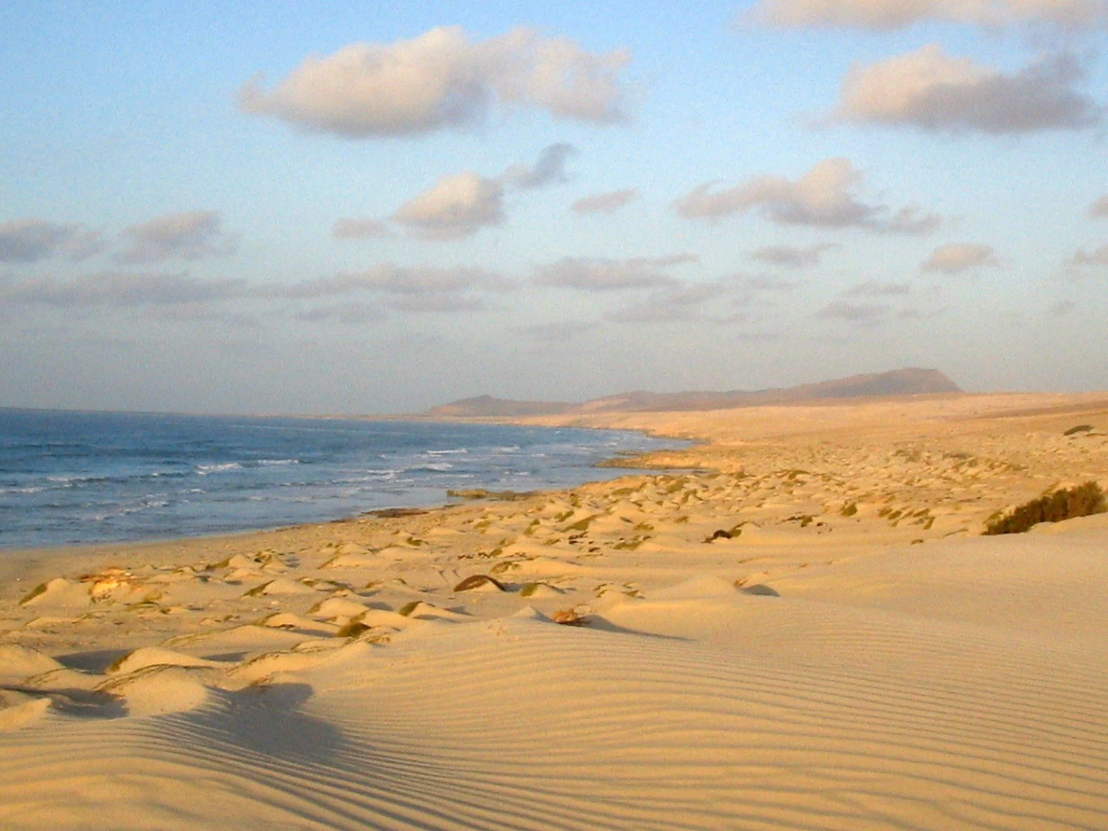 Spiaggia di sabbia dorata con onde che si infrangono, cielo con nuvole sparse e montagne lontane all'orizzonte.