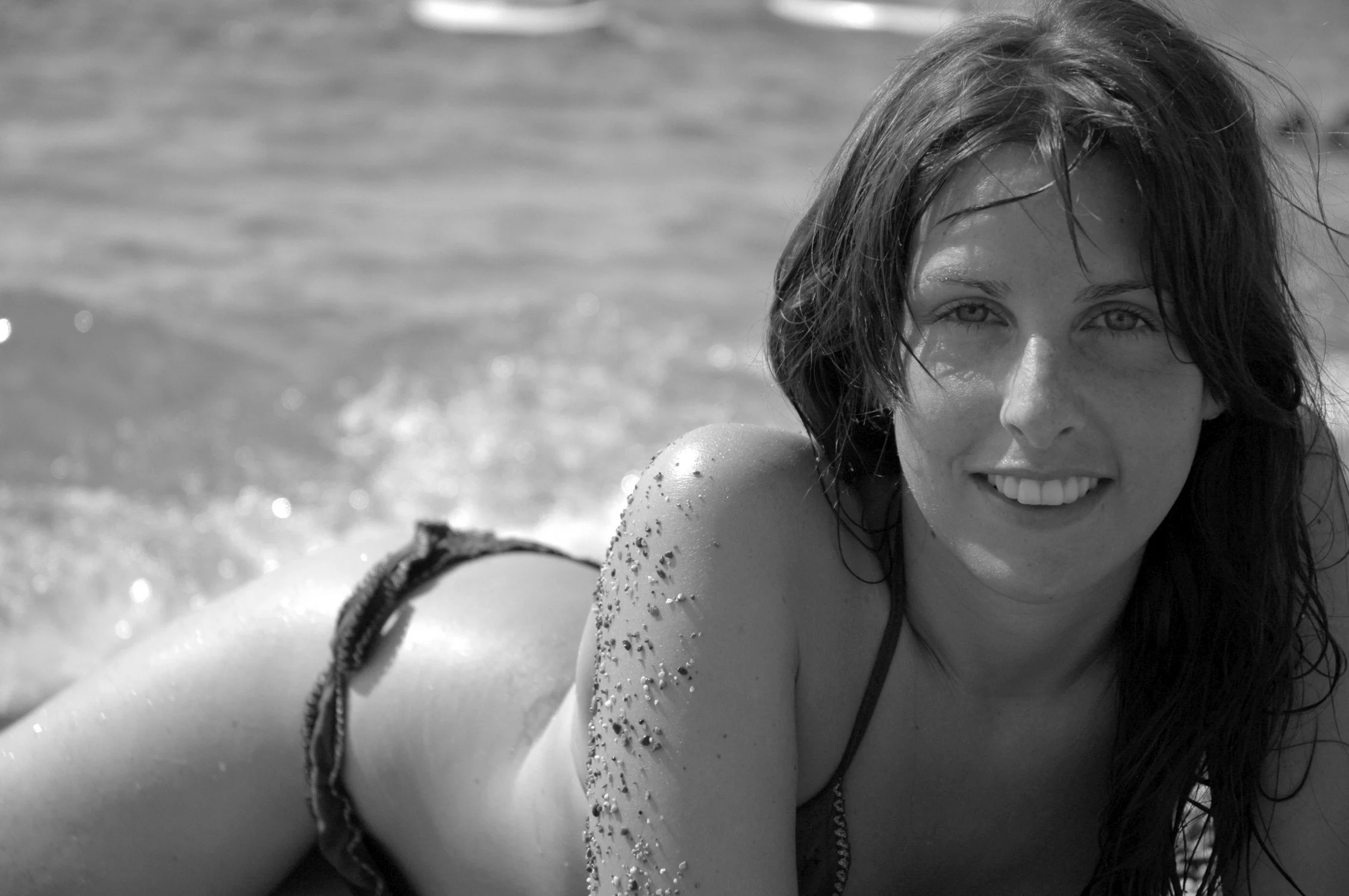 Ragazza sorridente con capelli umidi, che gioca in acqua, con brillante abbronzatura e dettagli di sabbia sulle spalle e braccia.