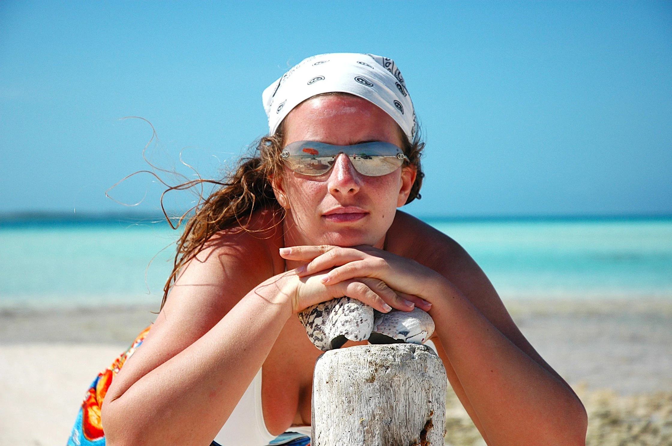 Femminile con cappello bianco, occhiali da sole riflettenti, rilassata appoggiata su una pietra vicino alla spiaggia, con il mare e il cielo azzurro sullo sfondo.