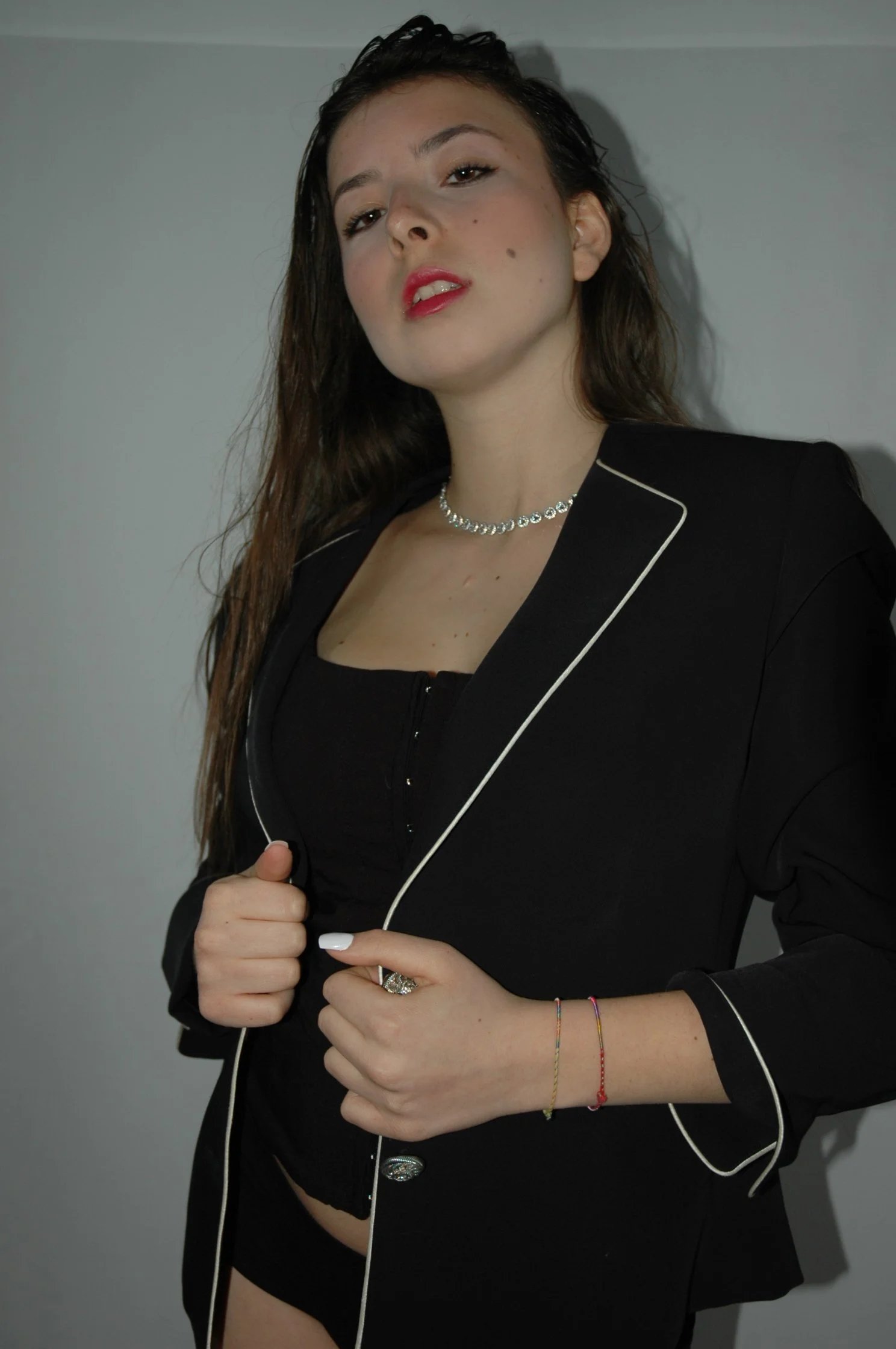 Ragazza con capelli castani ondulati e trucco rosato, indossa una giacca nera con piping bianco e una collana di diamanti, si trova di fronte a uno sfondo grigio chiaro.