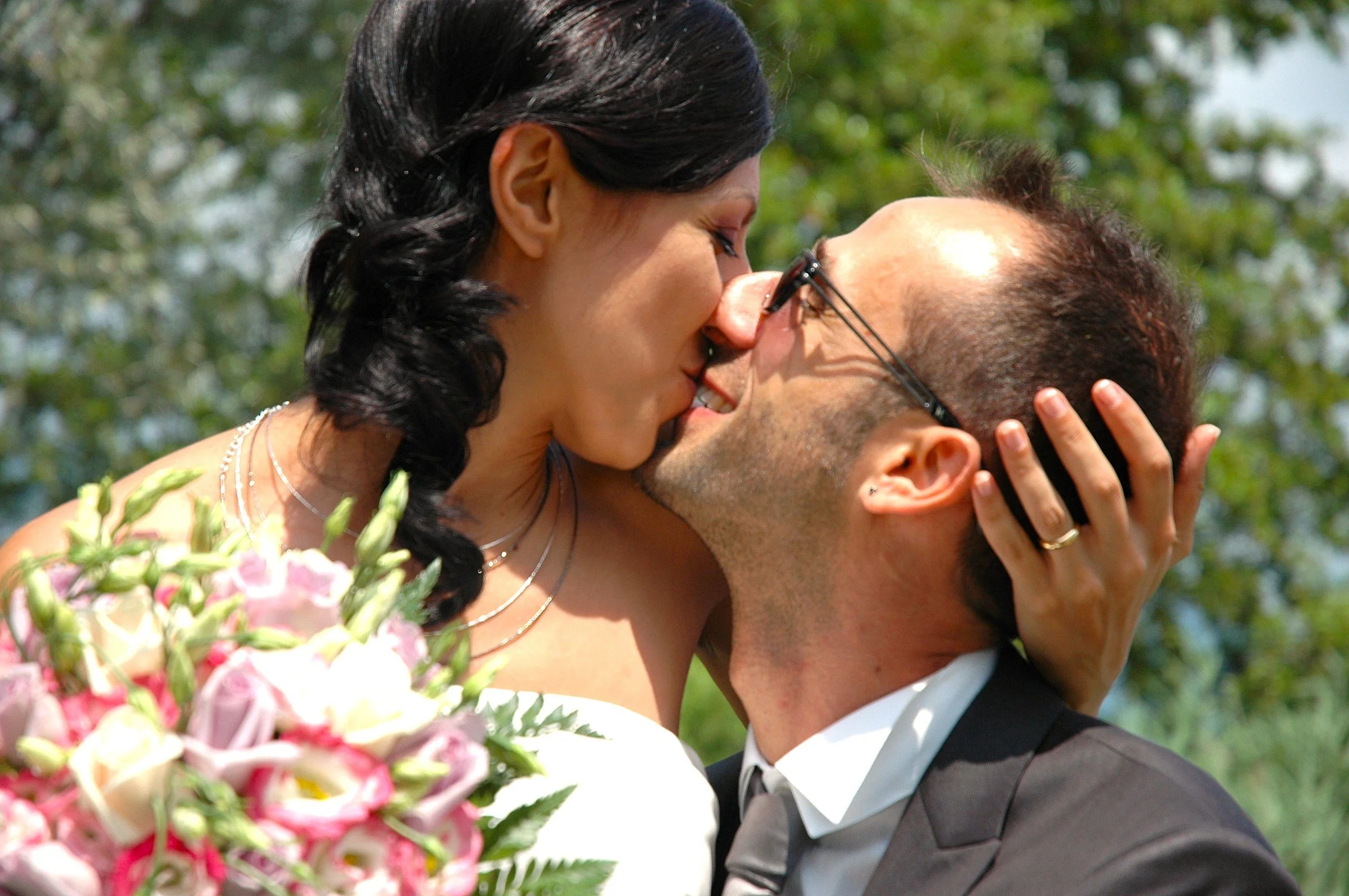 Una coppia di sposi si abbracciano felici in un matrimonio all'aperto, con sopra un mazzo di fiori rosa e bianchi.
