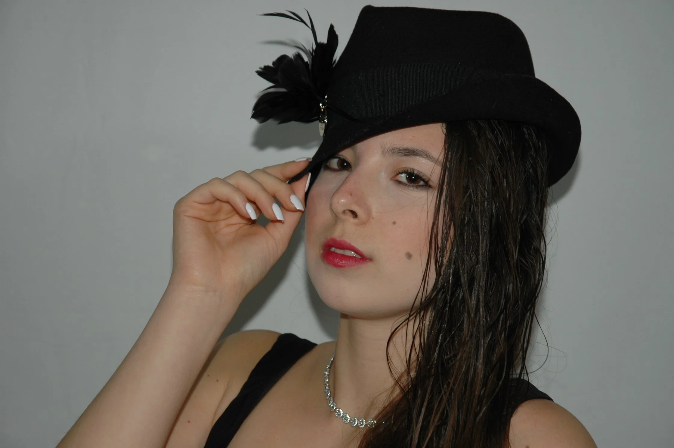 Una giovane donna con i capelli mossi indossa un cappello nero con piume e un collier di brillanti, sorride leggermente mentre tocca il cappello con la mano destra.