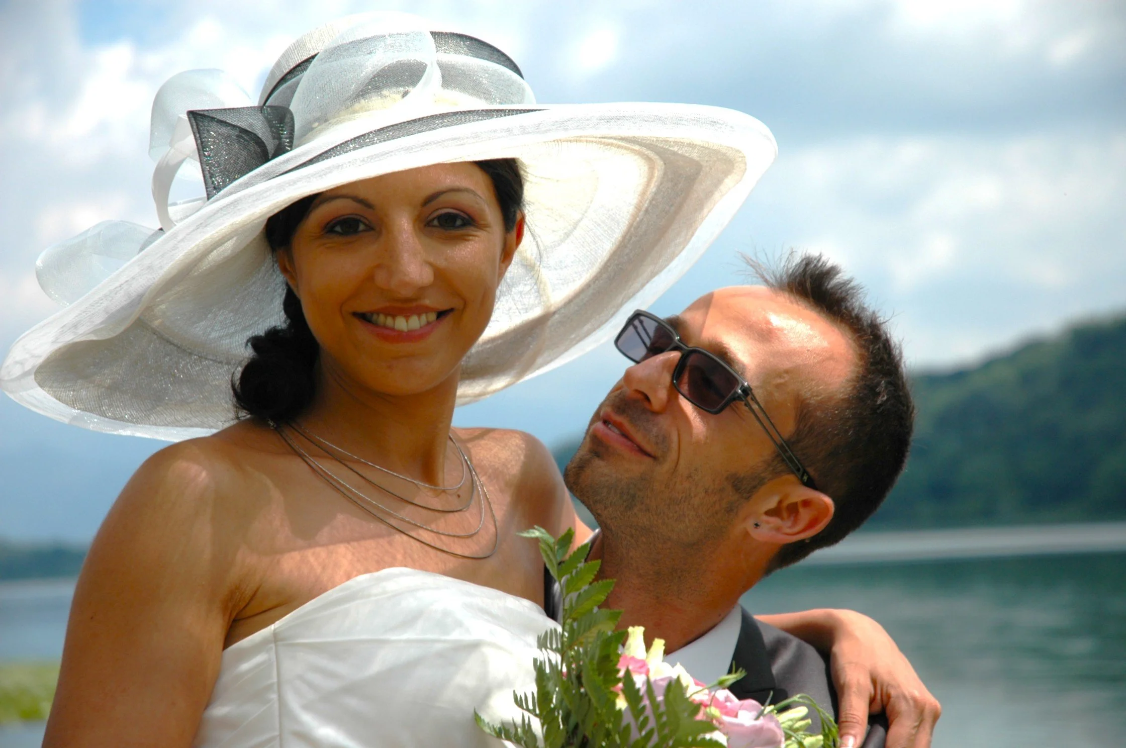 Una coppia di sposi al matrimonio sulla spiaggia, sorridenti, con la sposa che indossa un cappello bianco grande e il vestito bianco, e lo sposo in abito scuro con occhiali da sole.