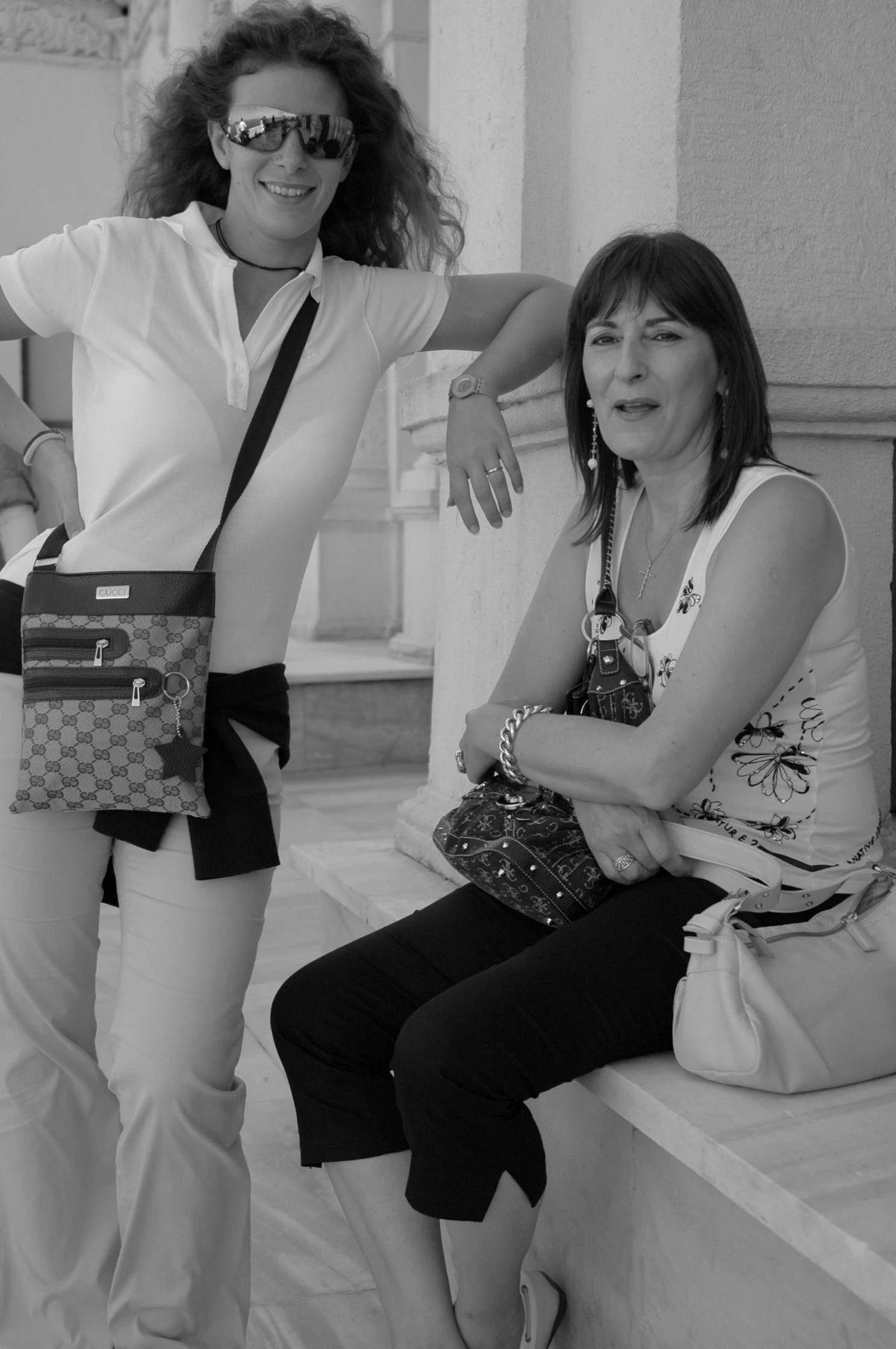Due donne sorridenti posano su una panchina, una di loro indossa grandi occhiali da sole e una camiseta bianca, l'altra indossa una maglietta con motivo floreale. Attraverso le loro espressioni sembra che siano felici e ci siano in un luogo pubblico.