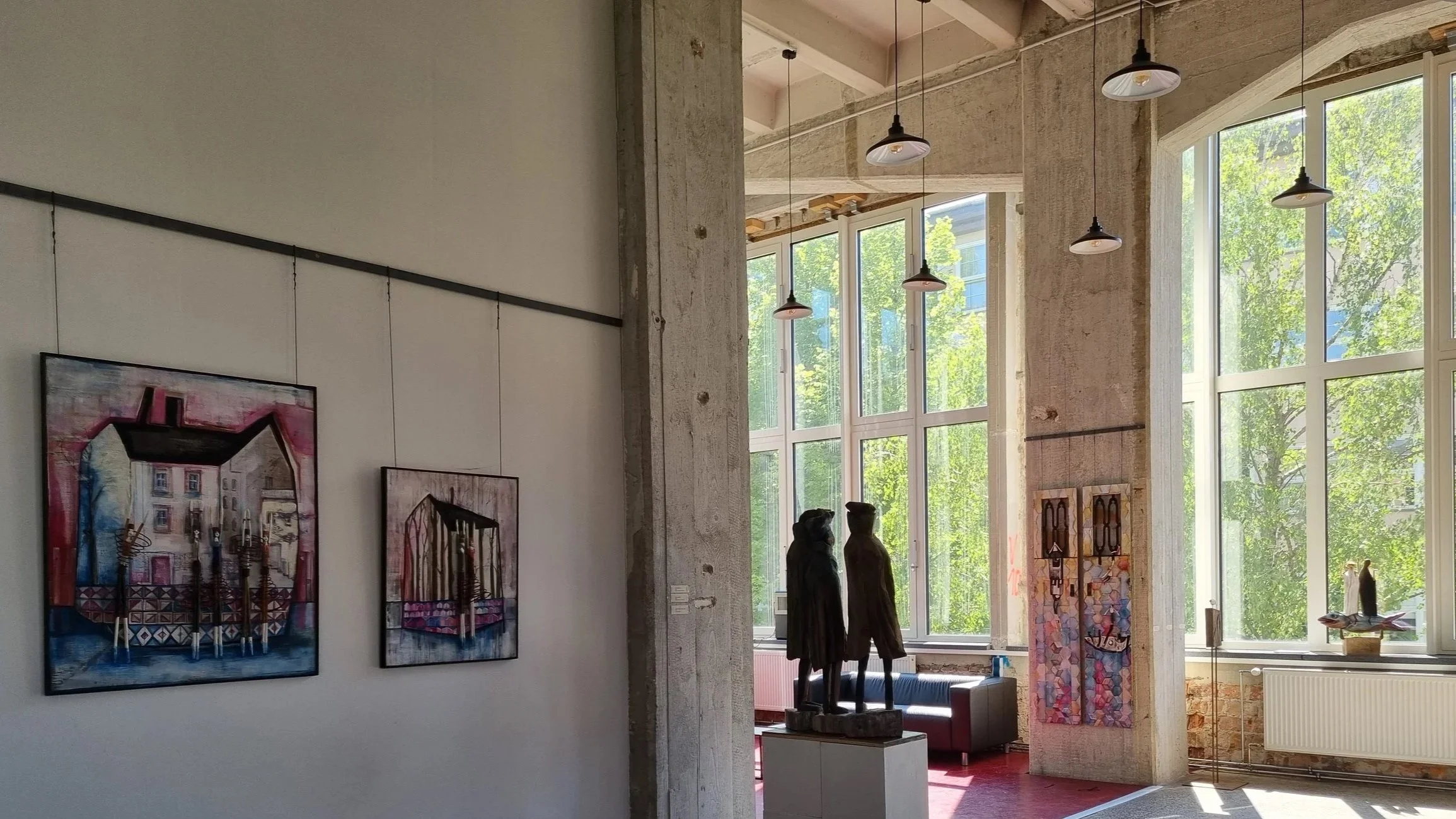 Innenaufnahme eines Kunstgalerieraums mit großen Fenstern, modernen Kunstwerken an den Wänden und Skulpturen.