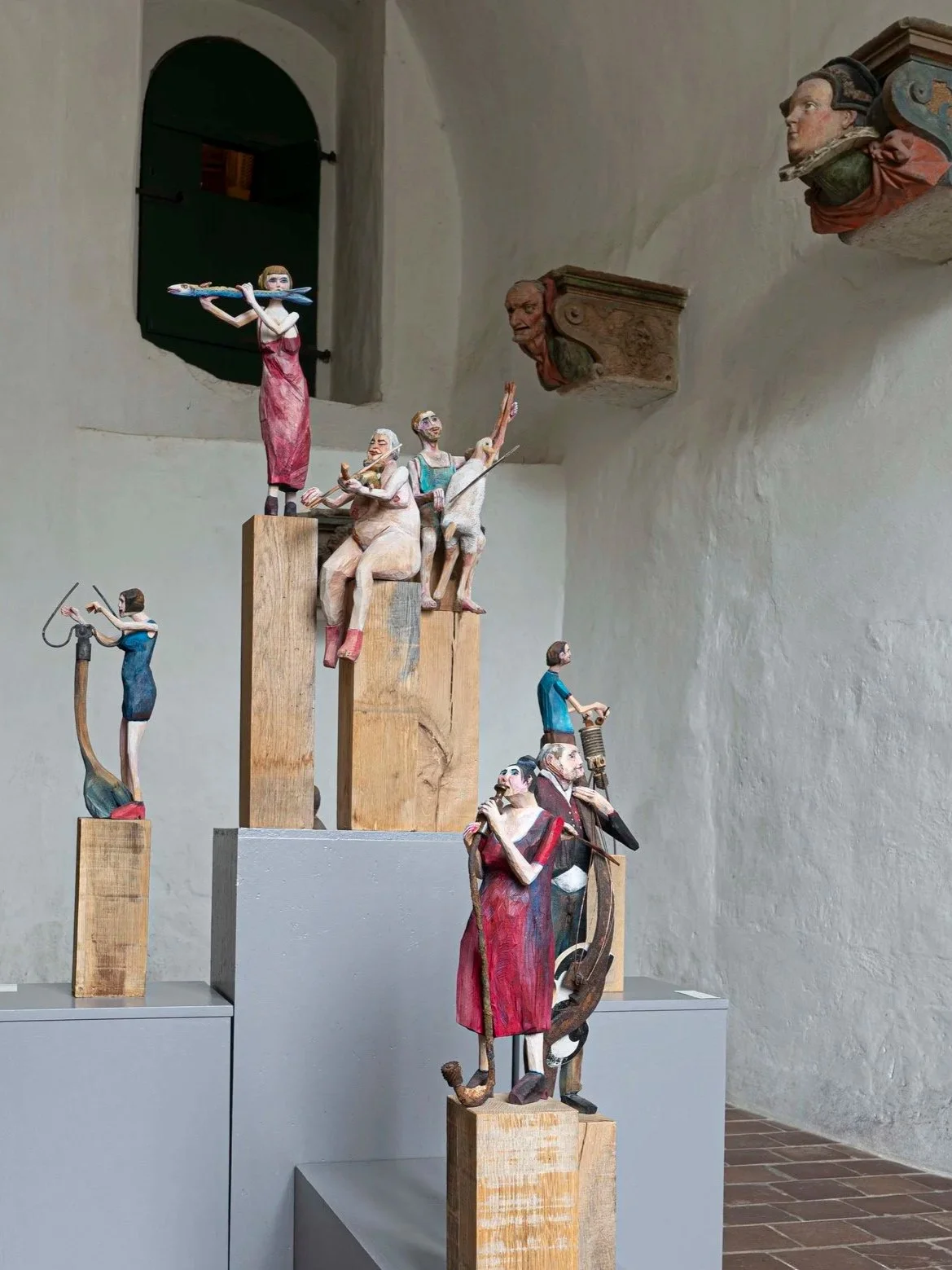 Holzskulpturen, die eine Gruppe von Musikern und Sängern darstellen, auf Holzsäulen in einem Raum mit weißen Wänden und Fenster. Es gibt auch Wandreliefs von Kopfsskulpturen.