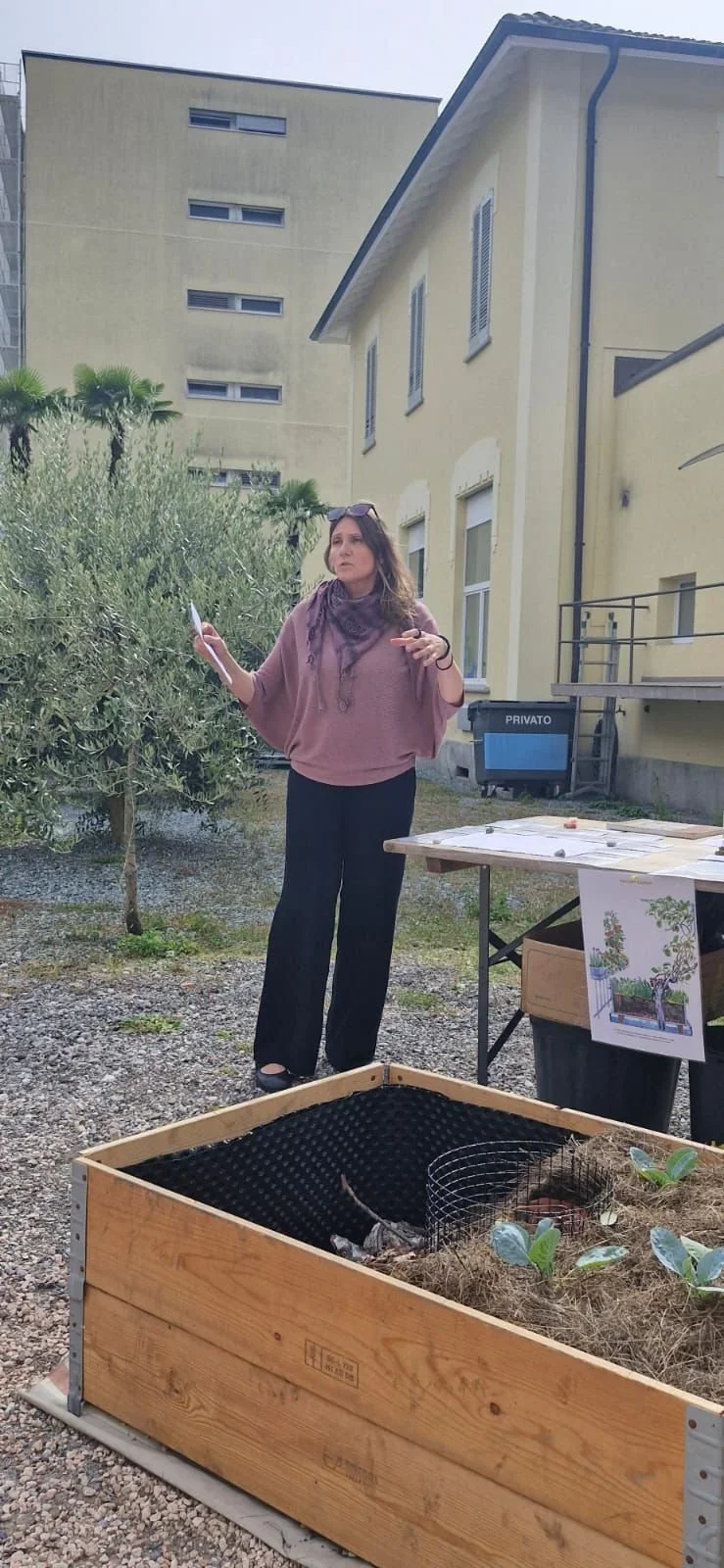 Woman explaining in an outdoor setting near a raised garden bed, holding a paper, with a table displaying gardening supplies and plants in the background.