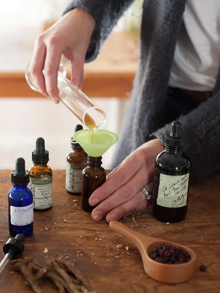 A woman measuring herbal extract with a funnel, surrounded by bottles, herbs, and a spoonful of dried herbs on a wooden table.