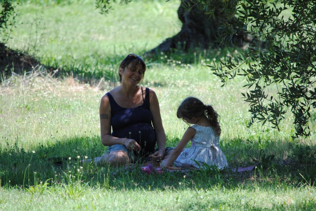 A woman and a young girl sitting on a grassy area under a tree, sharing a peaceful moment in nature.