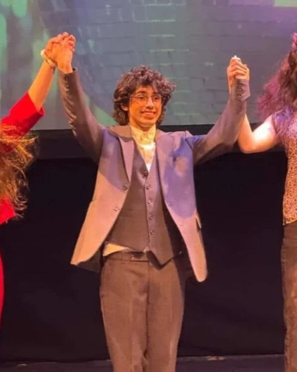 Ramiro, a young man with curly hair wearing a gray blazer, gray vest, and glasses, smiling and raising her hands during a stage performance.