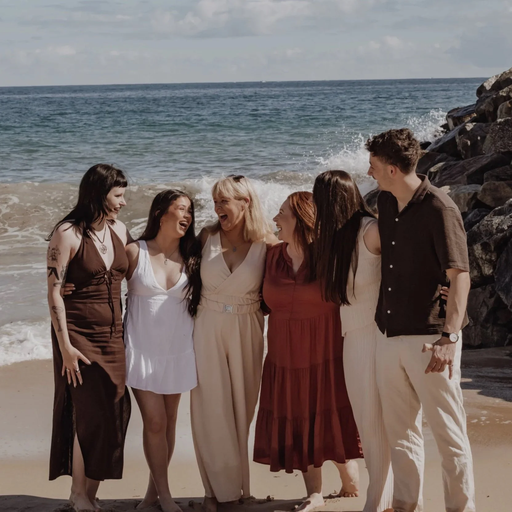 Six friends standing together on a beach, smiling and laughing, with ocean waves in the background.