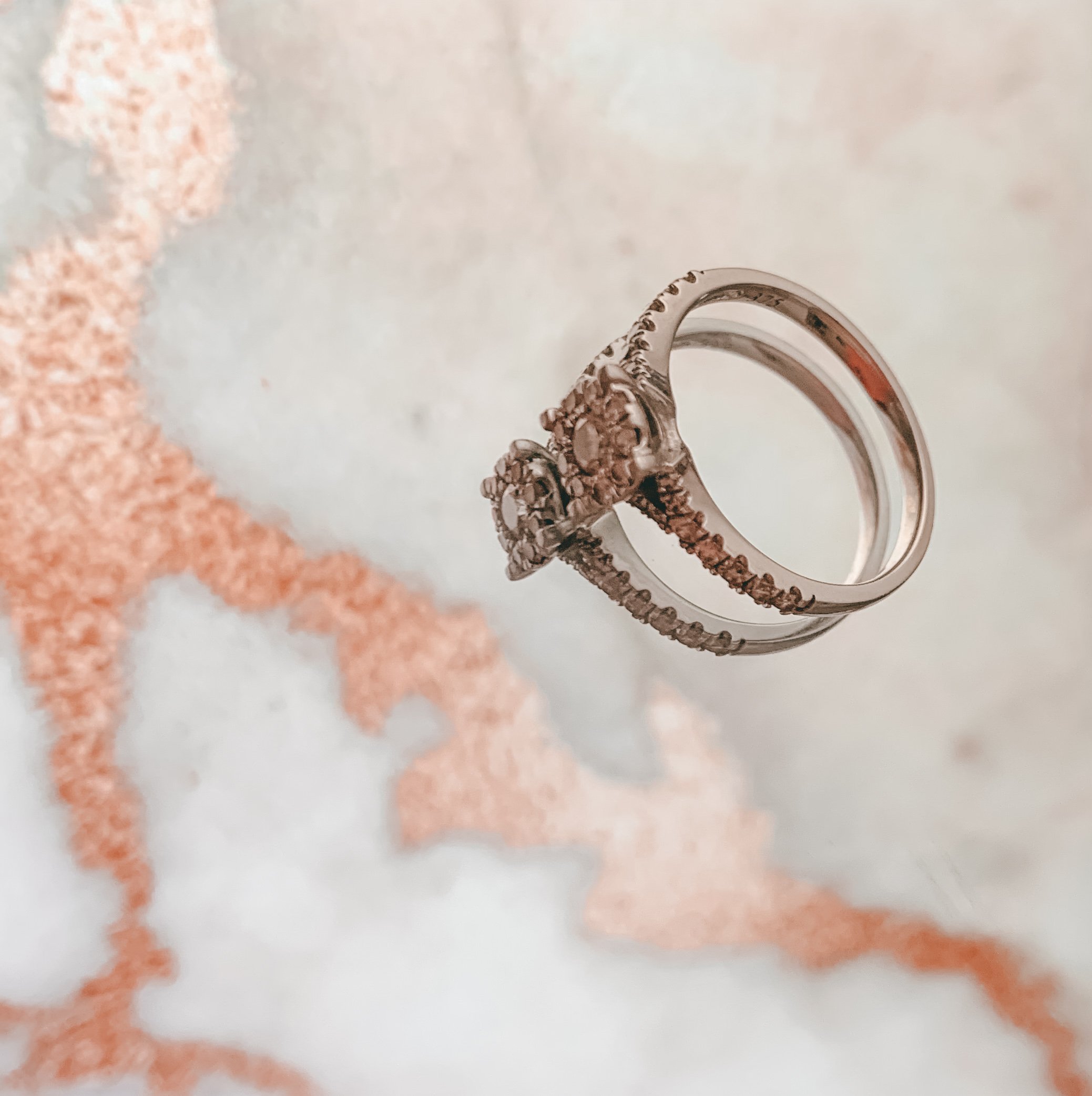 A close-up view of a silver ring with an intricate floral design, set against a blurred background of beige and ochre colors.