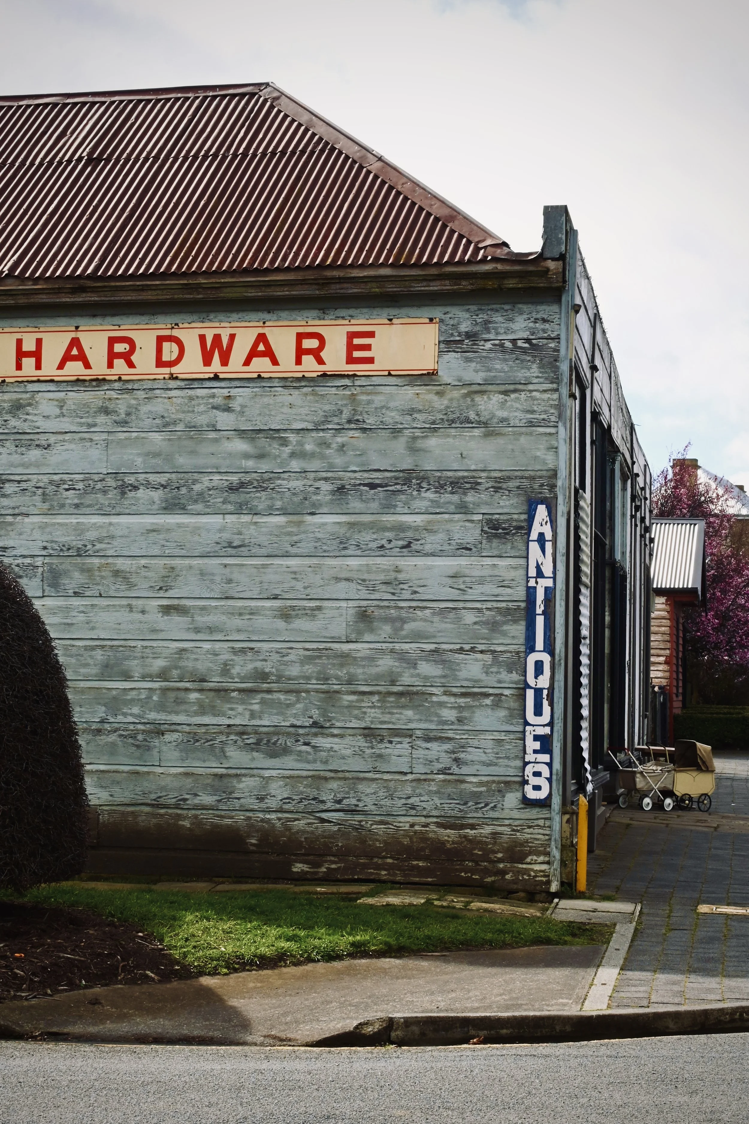 An exterior view of a vintage wooden hardware store with faded gray wood siding and a rusted red metal roof. A large sign with red letters spells 'HARDWARE'. A blue and white vertical sign on the right side reads 'SHOES'. There are some shop items li