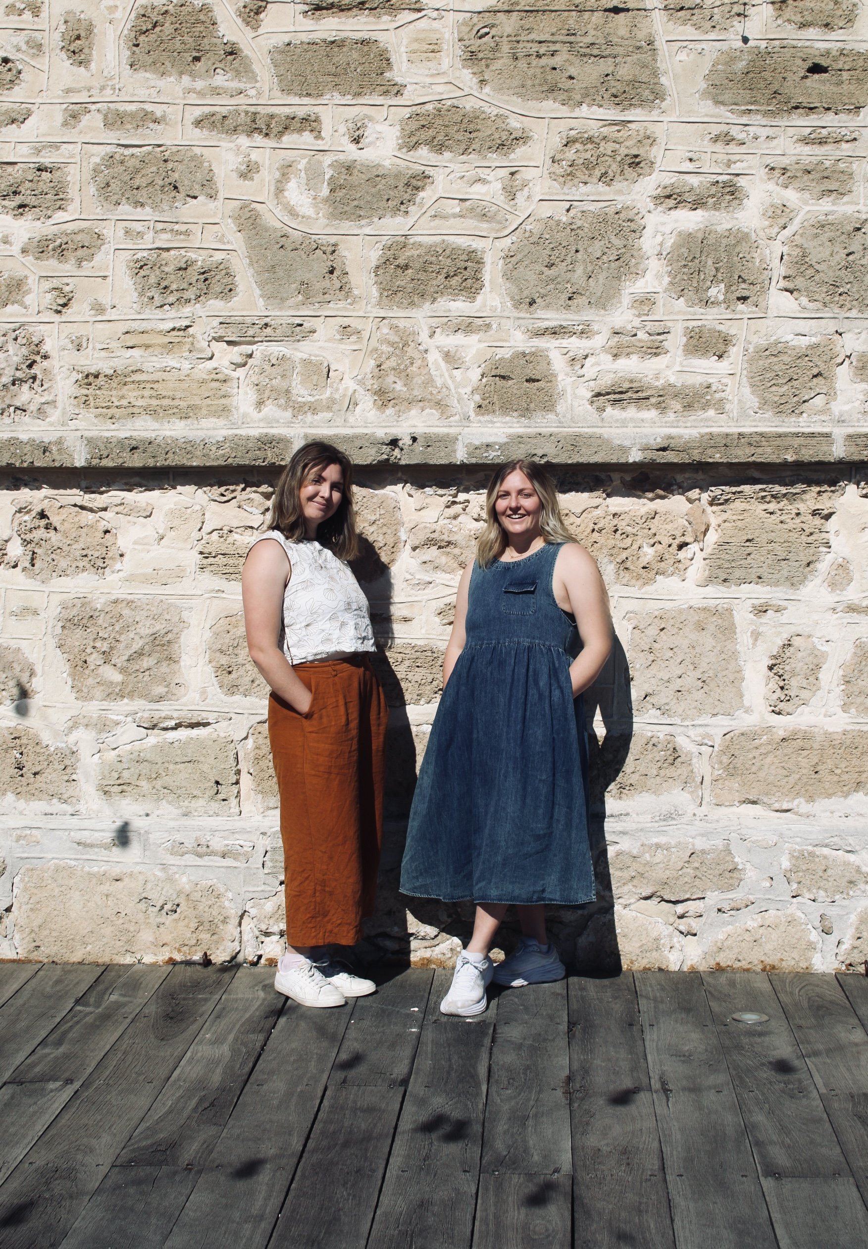 Two women standing side by side in front of a beige stone wall with large blocks. They are outdoors on a wooden deck, smiling at the camera, dressed casually.