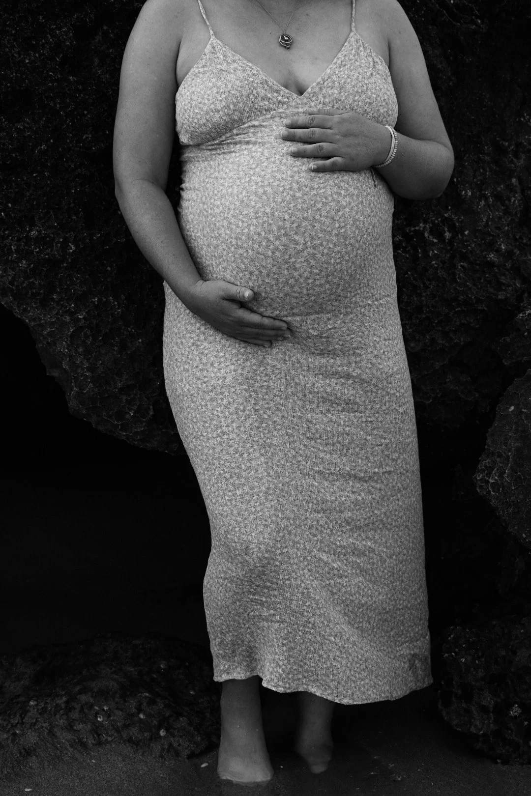A pregnant woman standing barefoot in front of rocks, wearing a long patterned dress and necklace with one hand on her belly and the other on her chest.