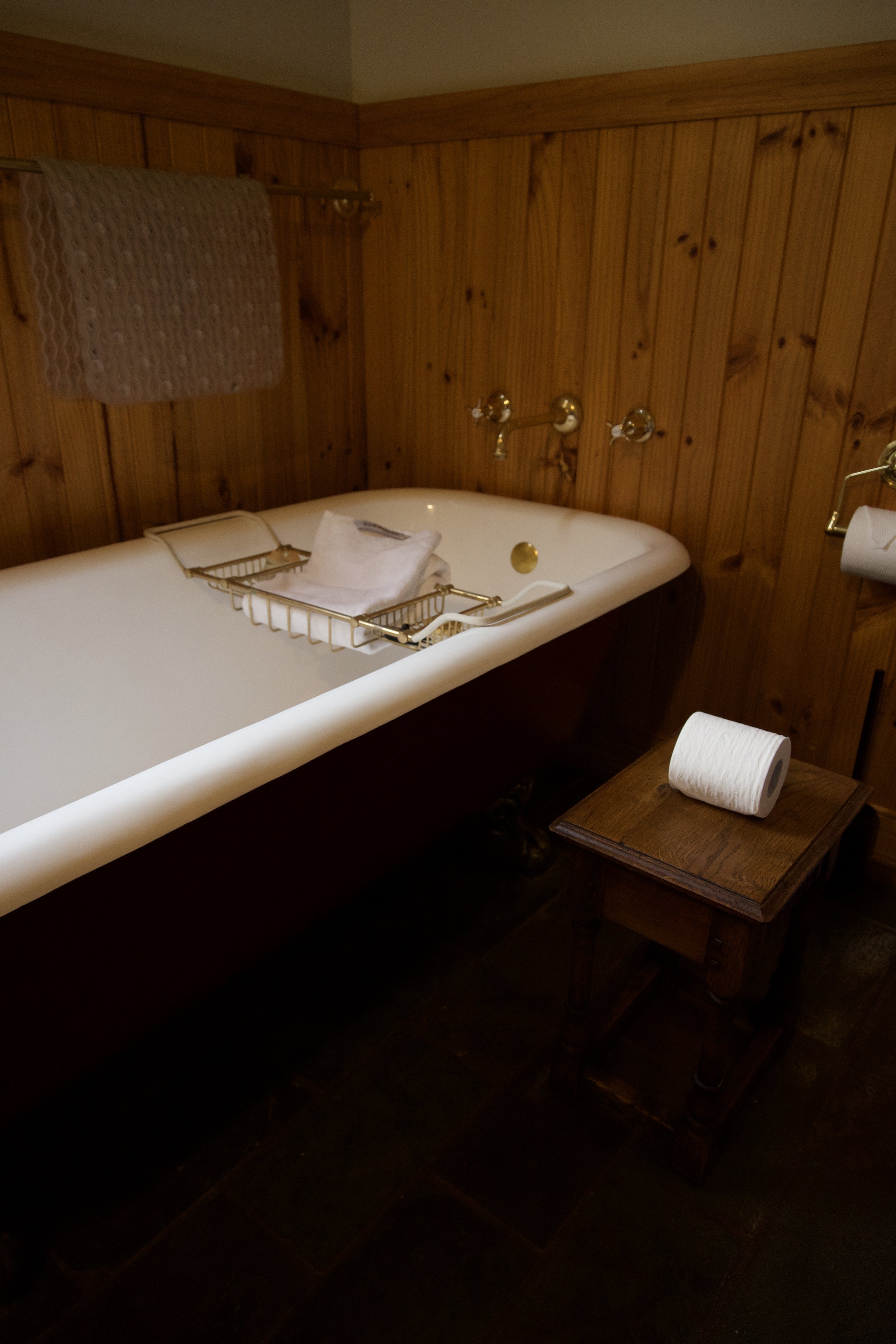 A vintage bathtub with a white interior, gold fixtures, and a wooden wall behind it. On the bathtub, there is a metal rack with folded white towels and a toothbrush. To the right of the bathtub, there is a small wooden table with a roll of toilet pap