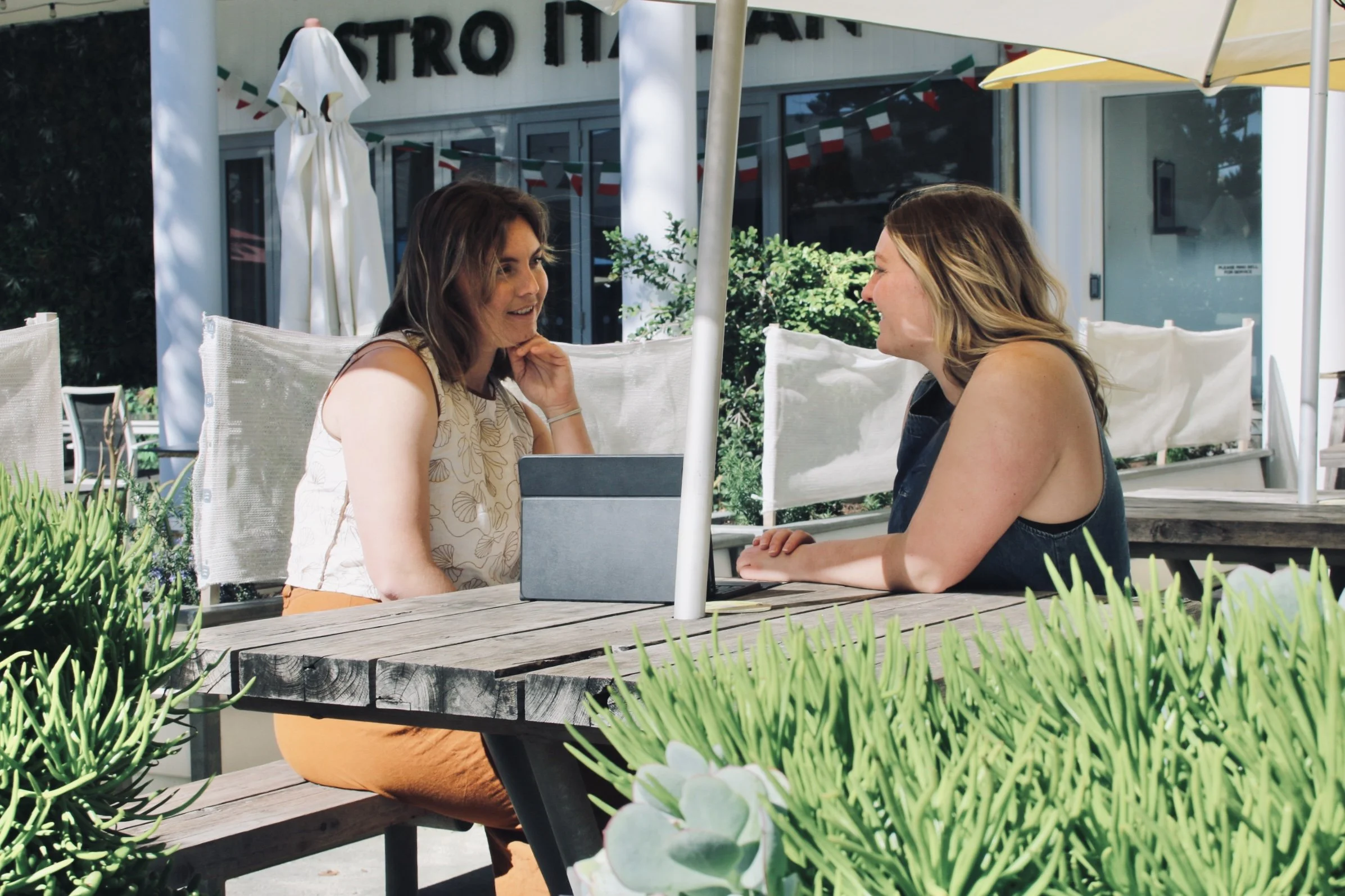 Two women sit at a table outdoors at a restaurant, engaging in conversation. One woman has brown hair and wears a sleeveless top, while the other has blonde hair and wears a black sleeveless top. There are large umbrellas providing shade, and greener