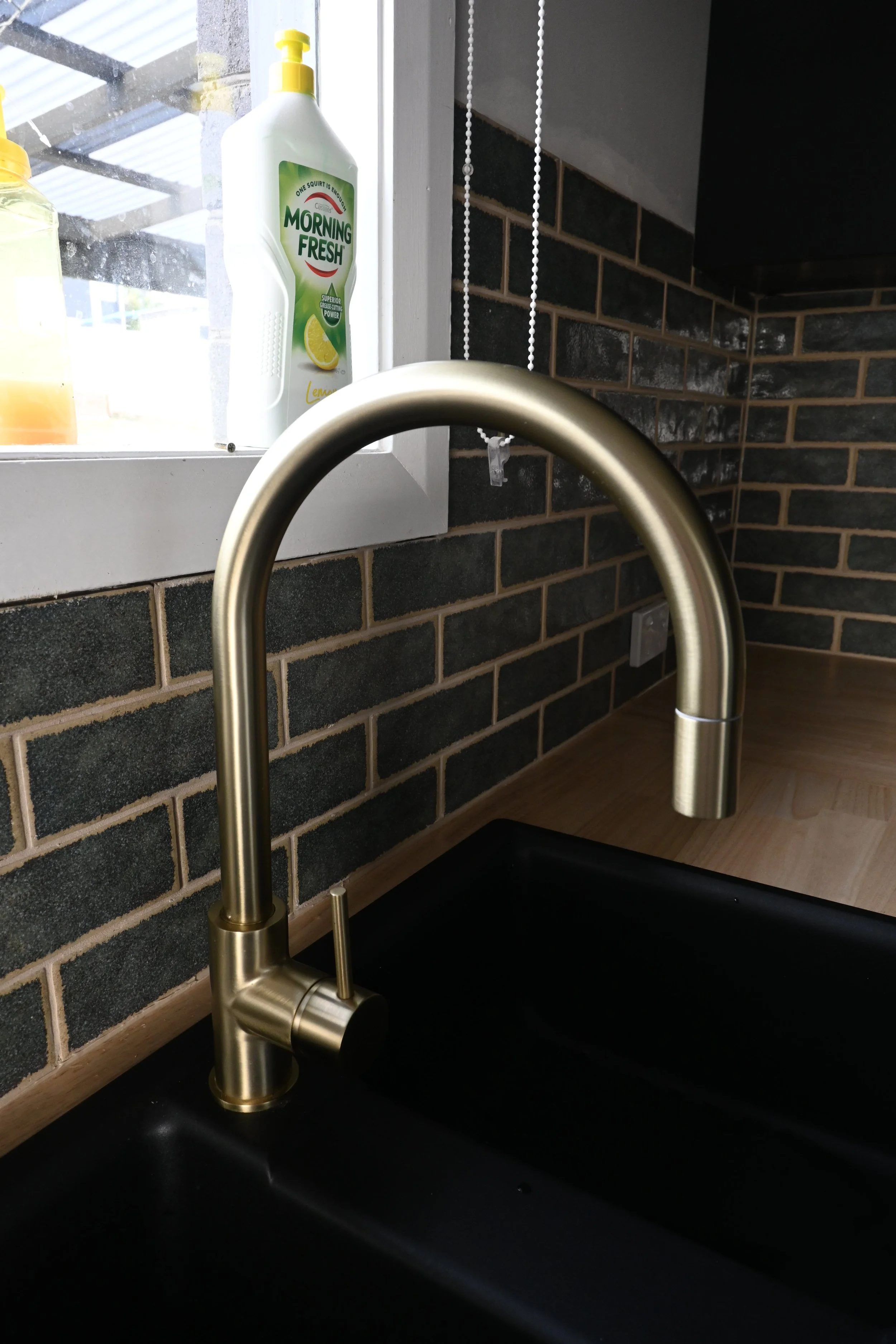 Close-up of a modern brass kitchen faucet over a black sink, with a brick backsplash and a window with a spray bottle of 'Morning Fresh' lemon dish soap visible in the background.