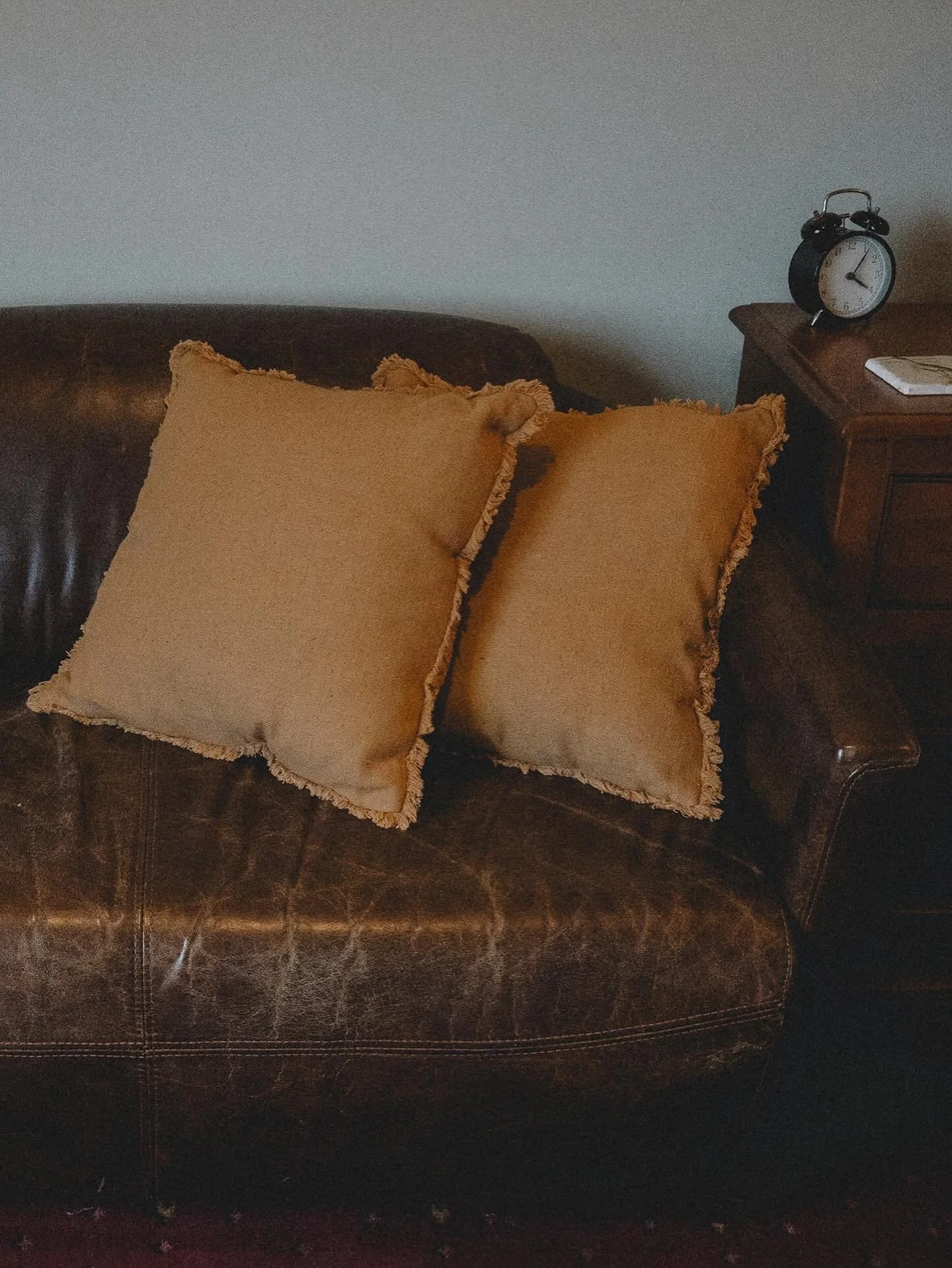 Two beige pillows with frayed edges on a dark leather sofa, with a wooden side table that holds a black alarm clock and a white item, against a plain wall.