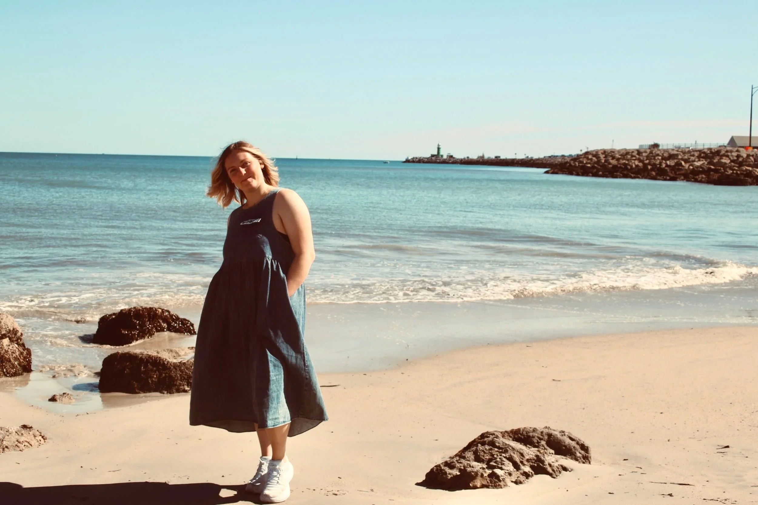 A woman with blonde hair wearing a sleeveless black dress and white sneakers standing on a Bathers Beach in Fremantle near rocks with ocean waves, a lighthouse, and a breakwater in the background.