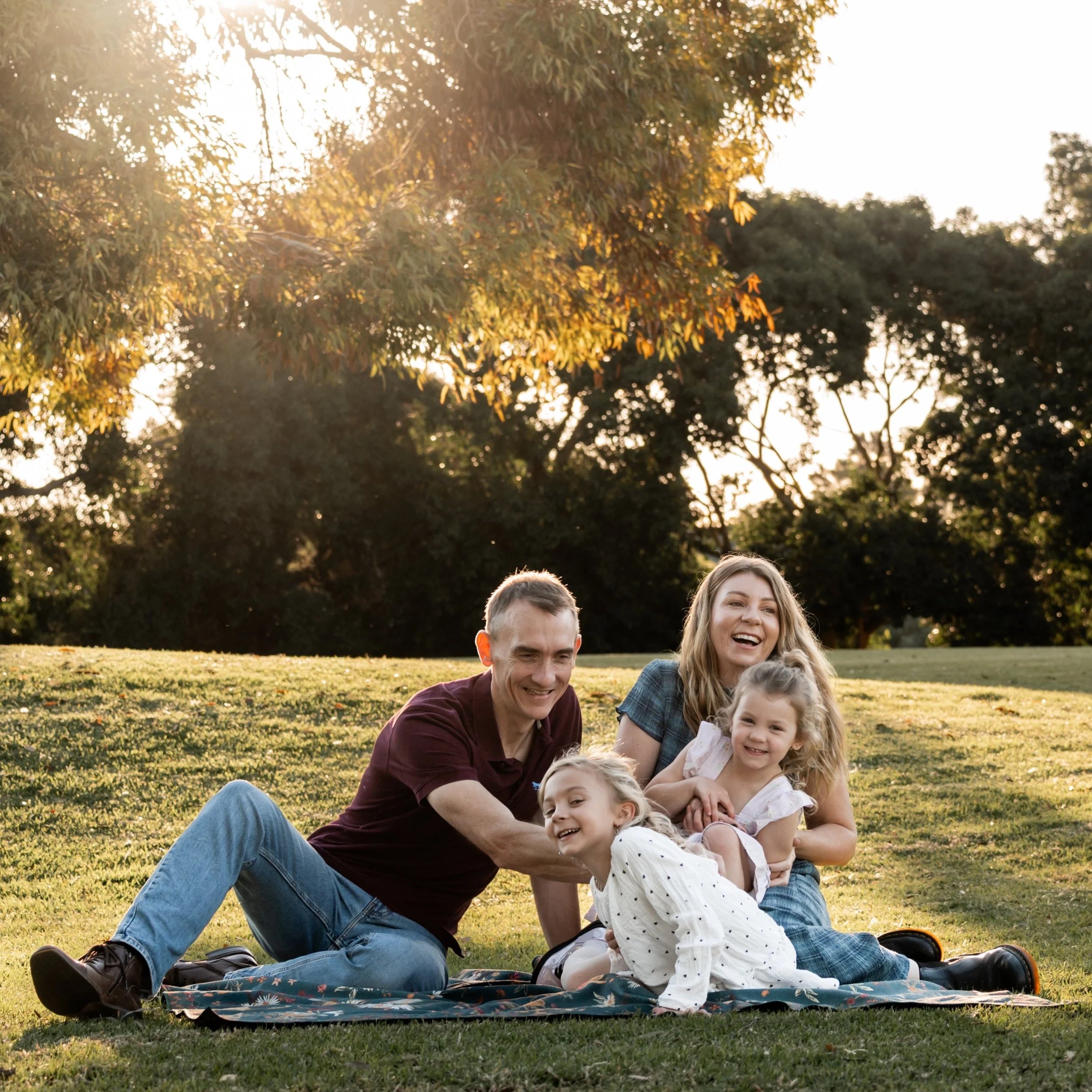 Family of four, with two adults and two children, sitting on a blanket in a park at sunset, smiling and enjoying the outdoor setting.