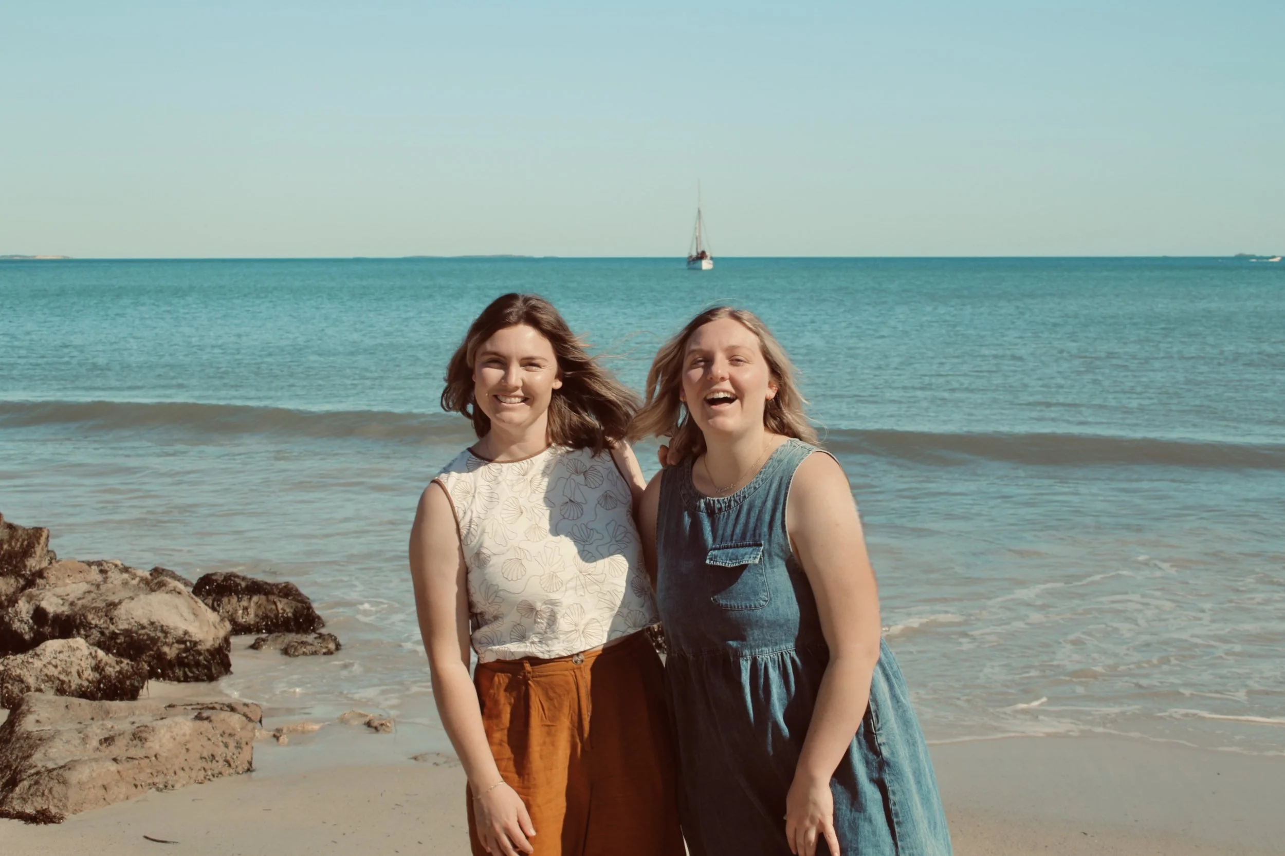 Two women smiling at the beach with ocean and sailboats in the background.