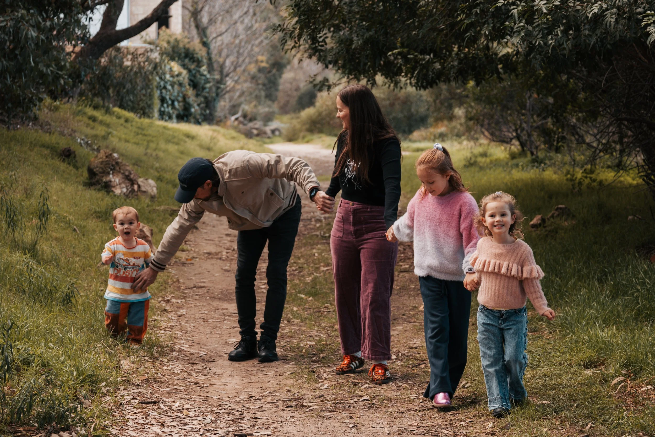 A family walking on a dirt trail in a green park with tall trees, with two women and children, one man bending down, holding hands and smiling.