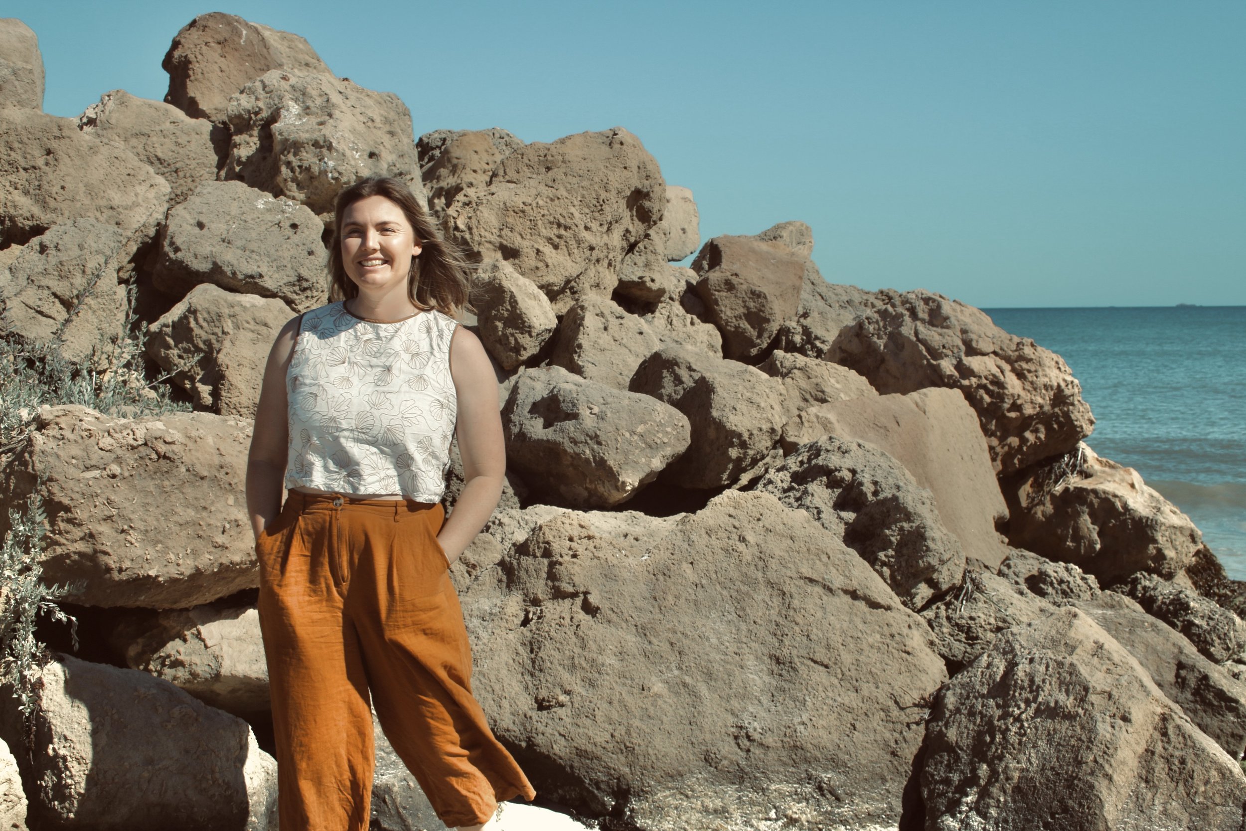 A woman smiling and standing in front of large rocks by the beach with the ocean and blue sky in the background.