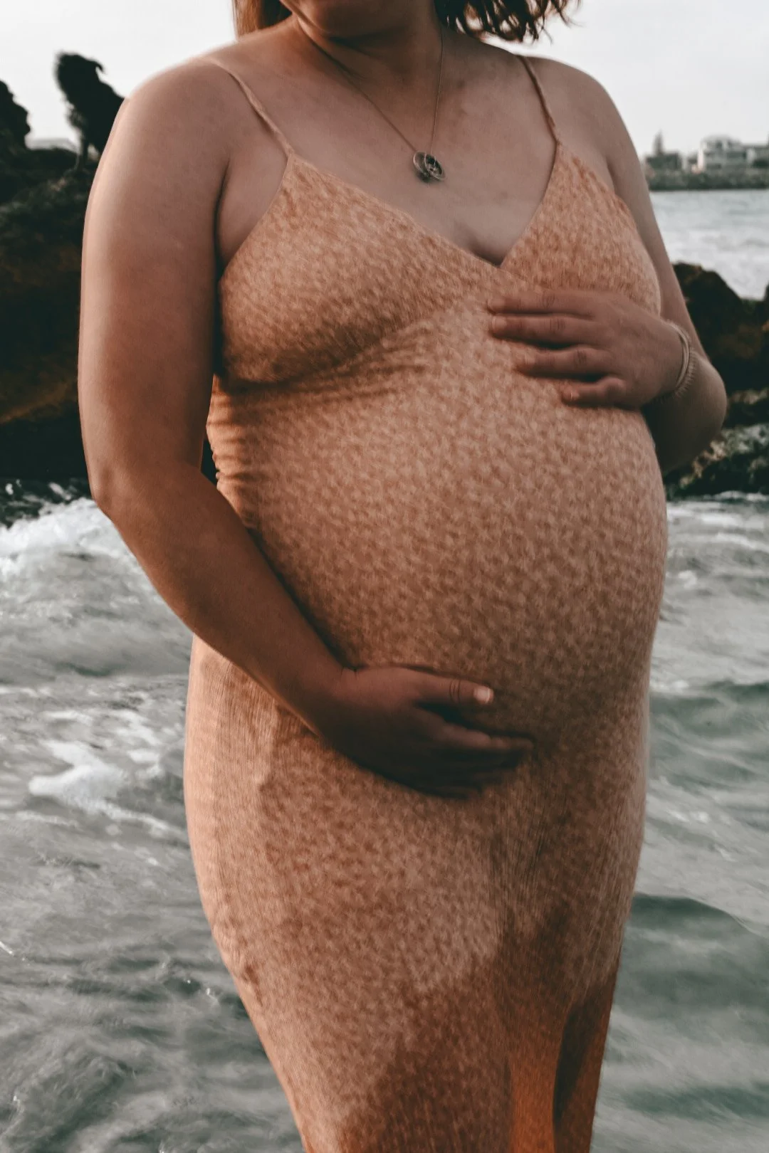 A pregnant woman in a peach dress standing by the ocean at North Coogee, holding her belly with one hand and resting her other hand on her chest.