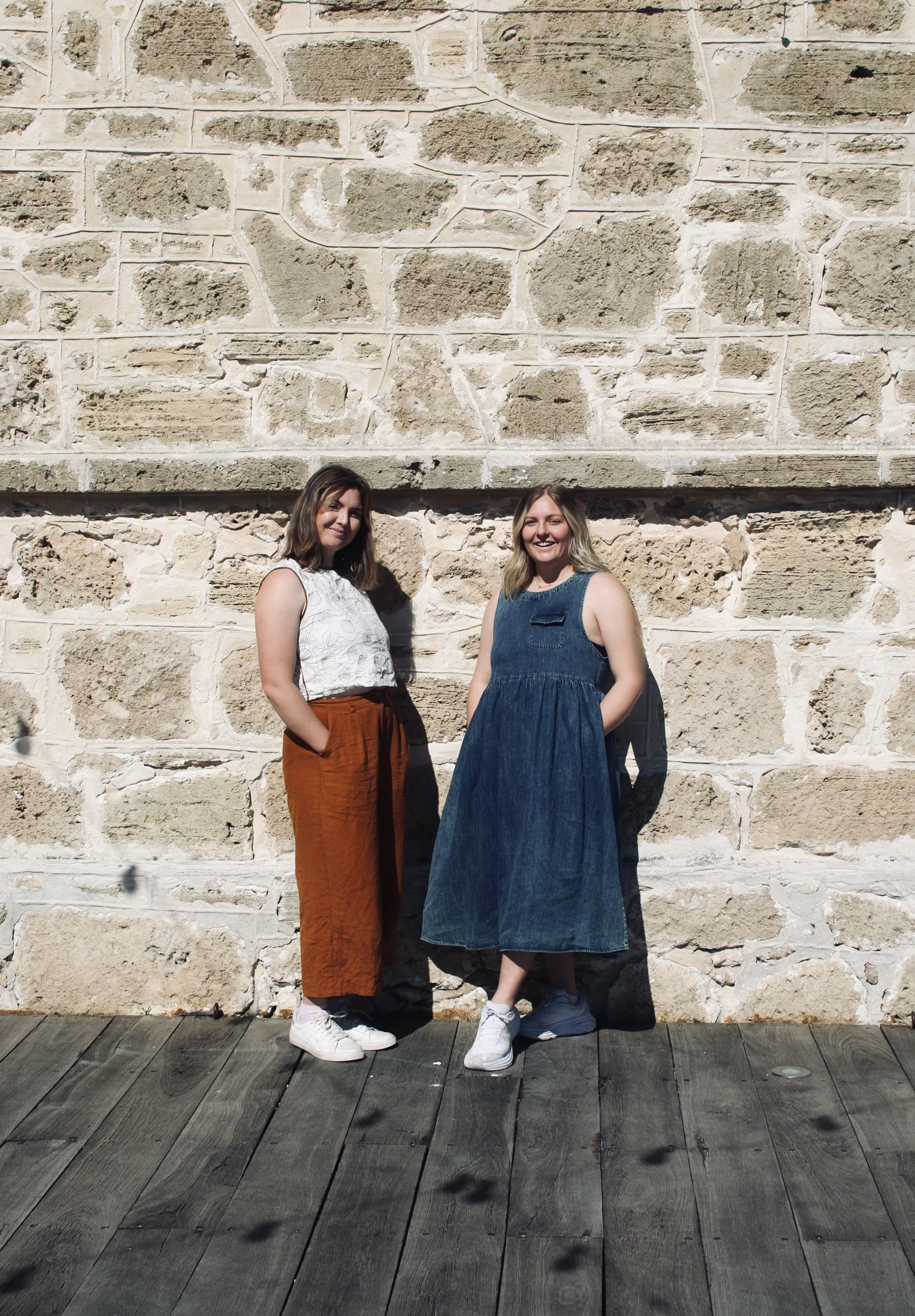 Two women standing in front of a stone wall, smiling at the camera. One woman is wearing a white top and orange pants, the other is wearing a blue dress. They are standing on wooden flooring.