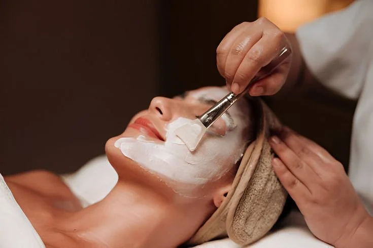Une femme reçoit un soin du visage dans un spa, avec un professionnel qui applique un masque facial blanc à l'aide d'une spatule.