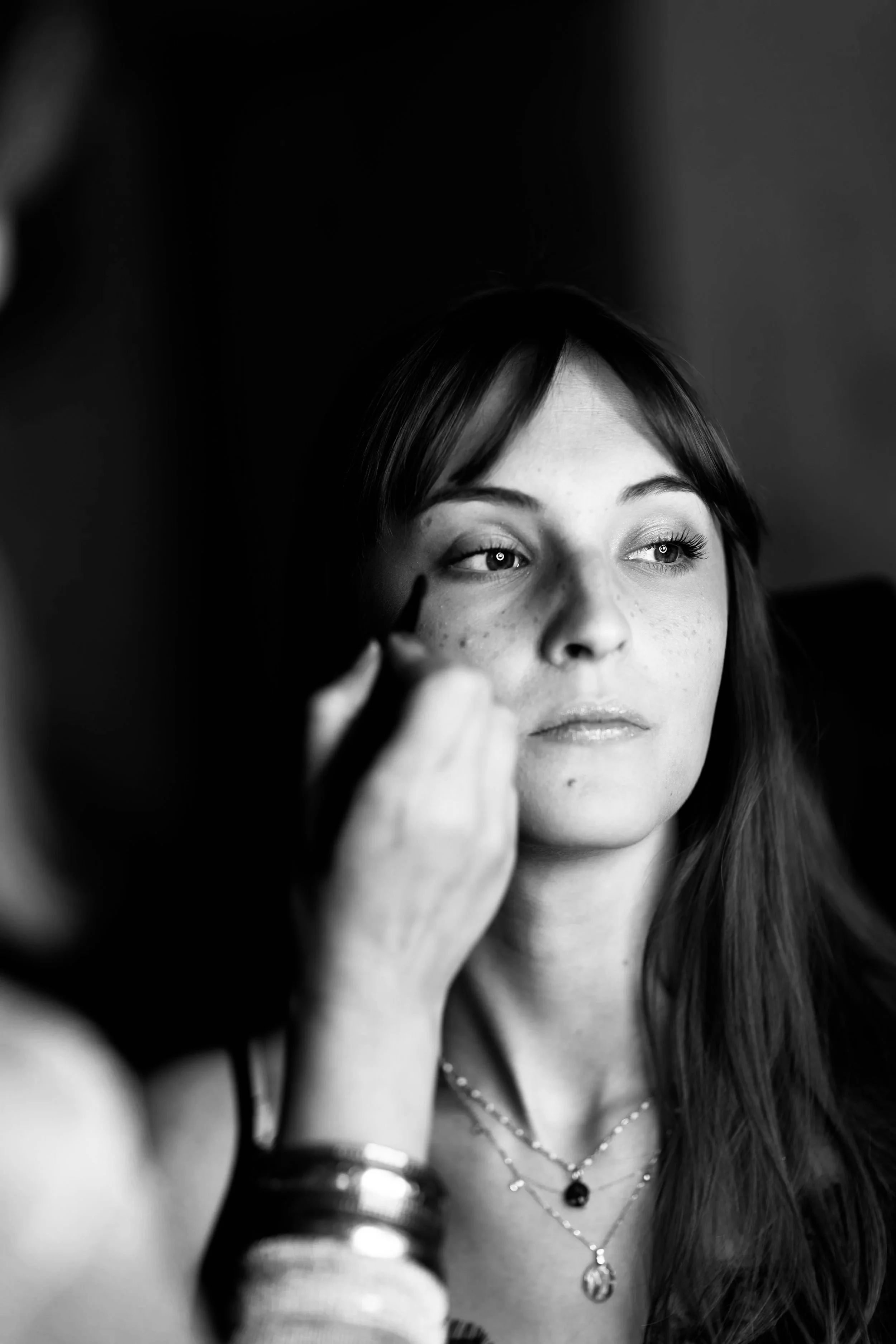 Une jeune femme aux yeux clairs et longues cheveux, maquillée, portant plusieurs colliers, se faisant maquiller ou s'appliquant un maquillage des yeux par une autre personne.