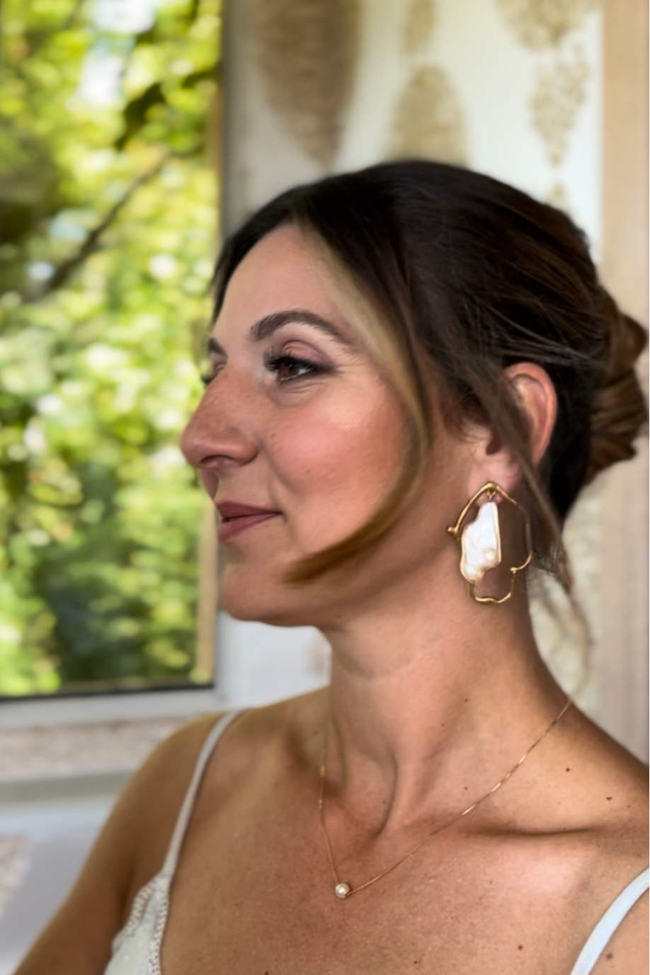 Portrait d'une femme avec une boucle d'oreille en forme de large pierre blanche et une chaîne fine avec une perle à l'avant, portant un maquillage subtil et un haut blanc fin, avec un fond naturel intégré par une fenêtre.