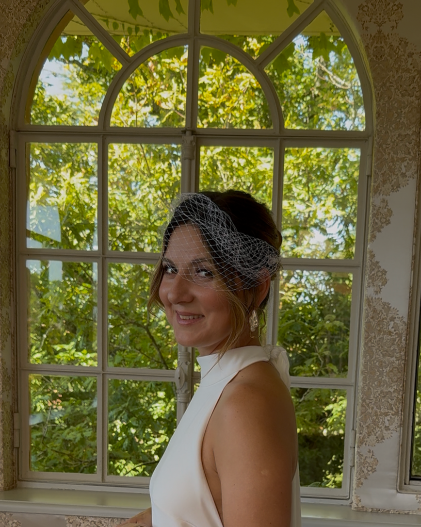 Une femme souriante portant une robe blanche et un voile, posant devant une grande fenêtre avec vue sur des arbres verts.