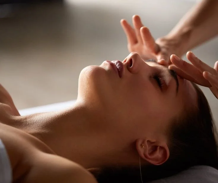 Une femme reçoit un massage, allongée sur le dos, avec un thérapeute qui utilise ses mains pour masser son visage ou son front.